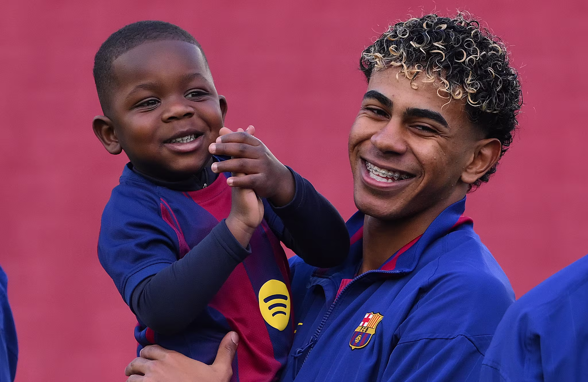 Lamine Yamal in a blue FC Barcelona jacket holds his brother Kenye in a matching jersey, both looking happy.