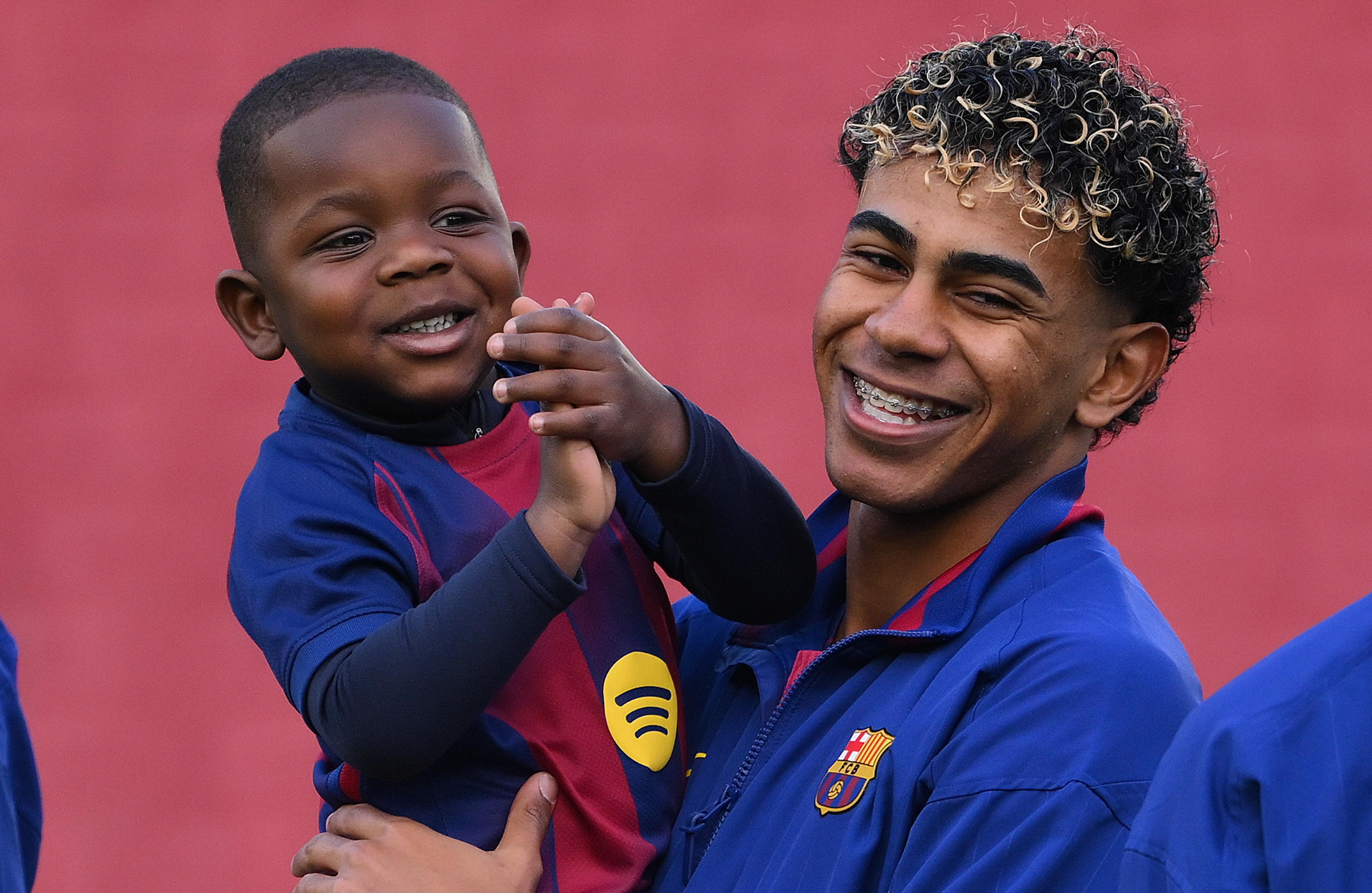 Lamine Yamal in a blue FC Barcelona jacket holds his brother Kenye in a matching jersey, both looking happy.