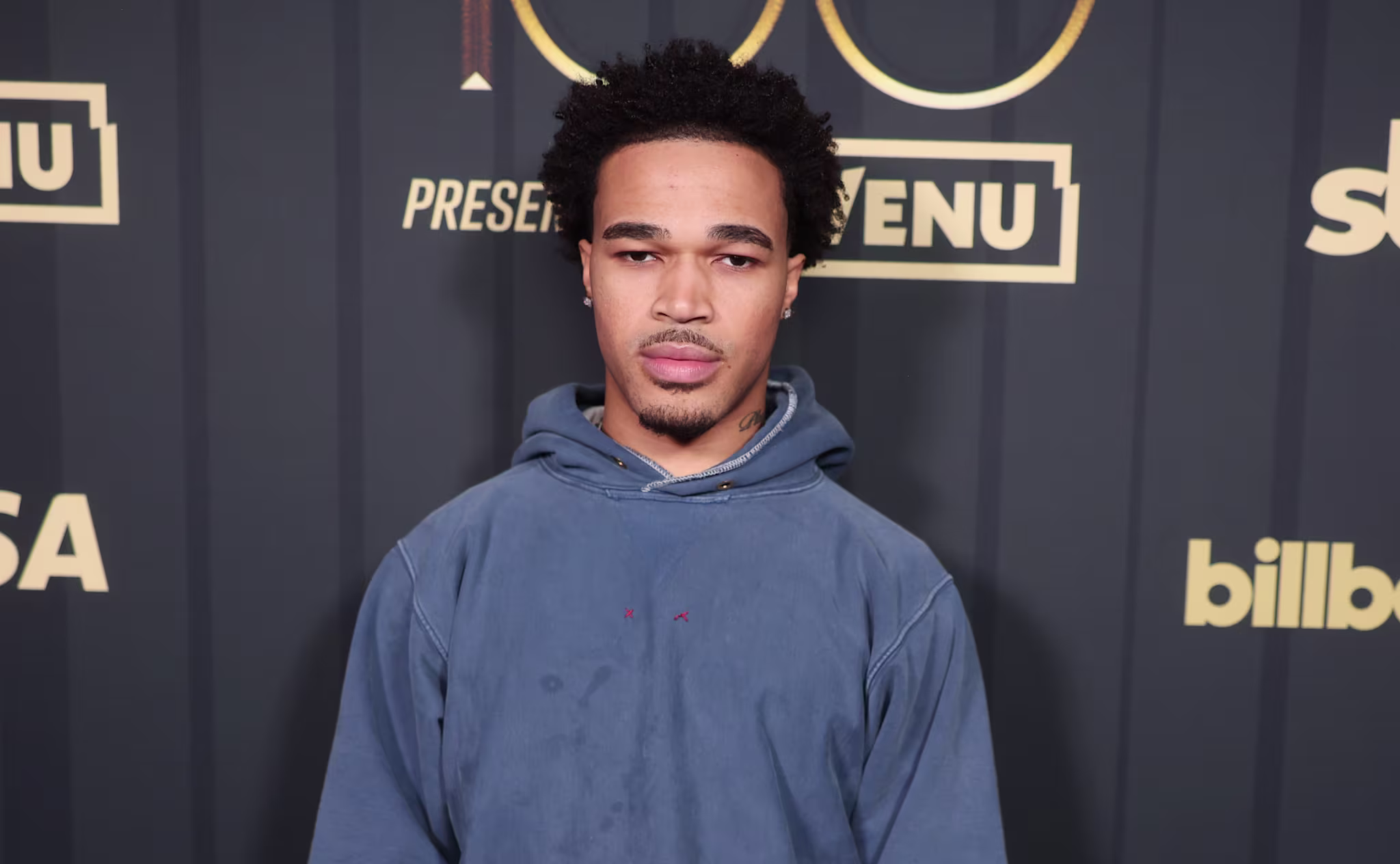 Plaqueboymax in a blue hoodie stands in front of a backdrop with logos like VISA, Billboard, and sbe.