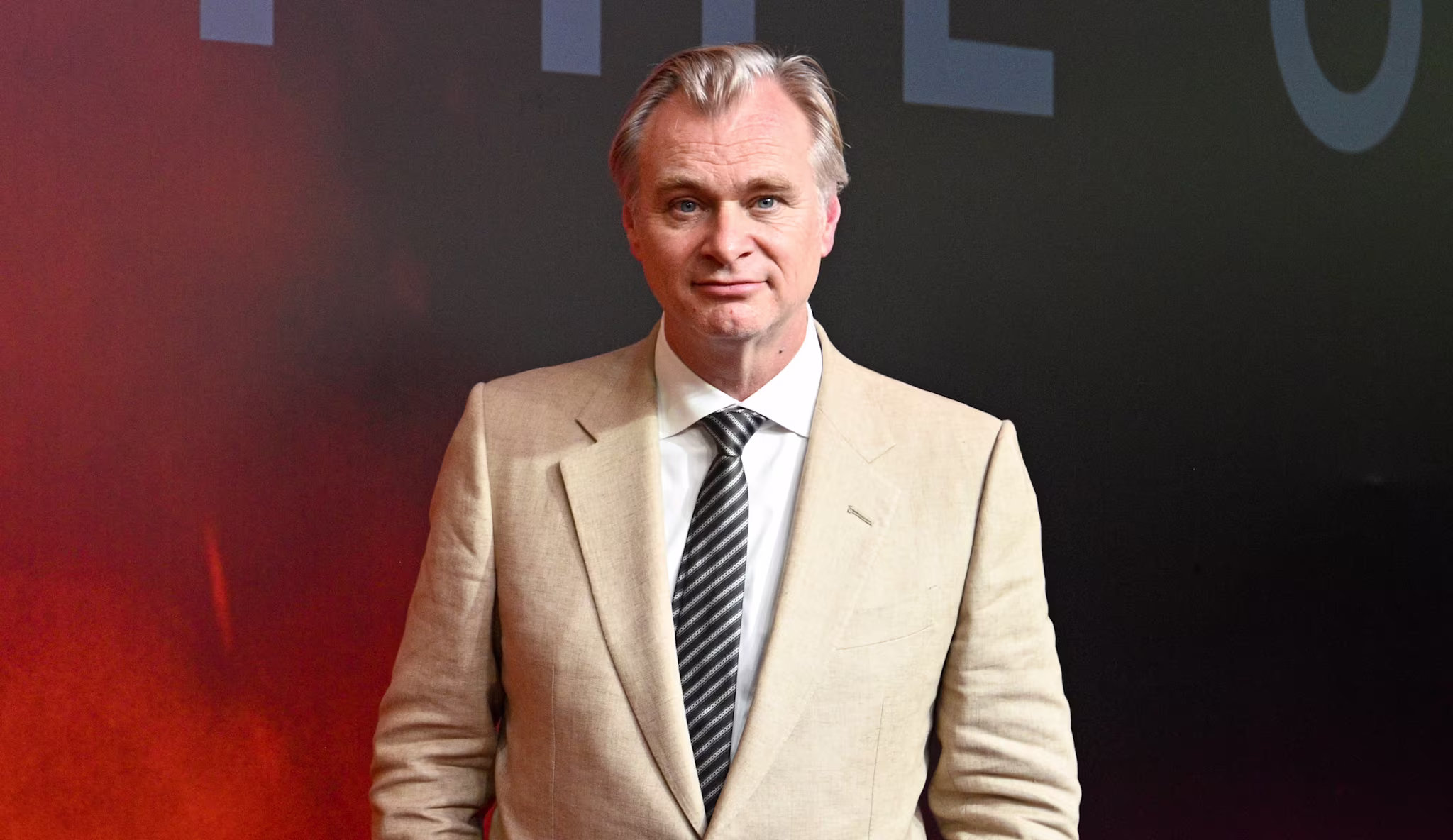 Christopher Nolan in a beige suit with a striped tie stands on a red carpet with a dark background.