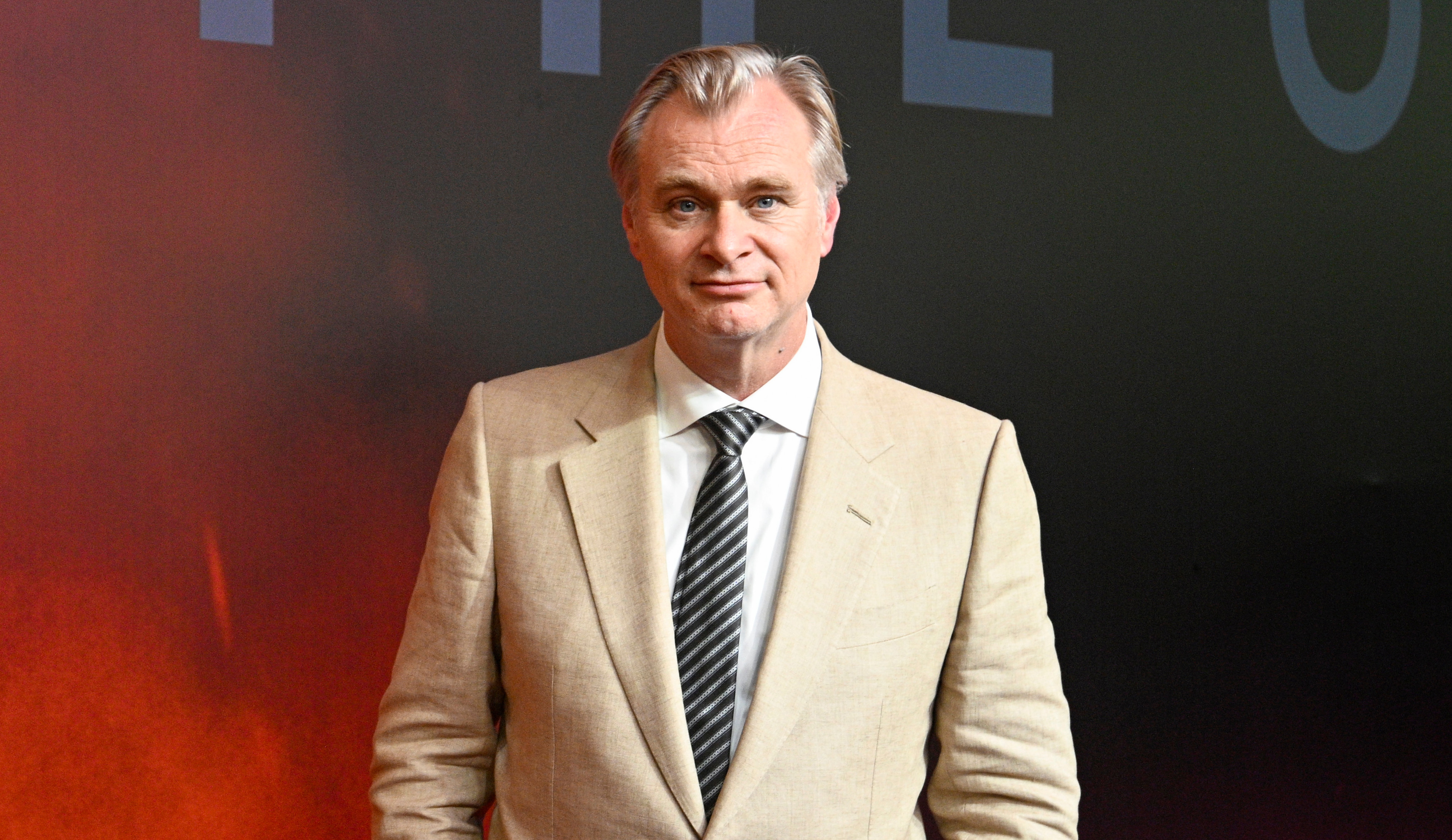 Christopher Nolan in a beige suit with a striped tie stands on a red carpet with a dark background.