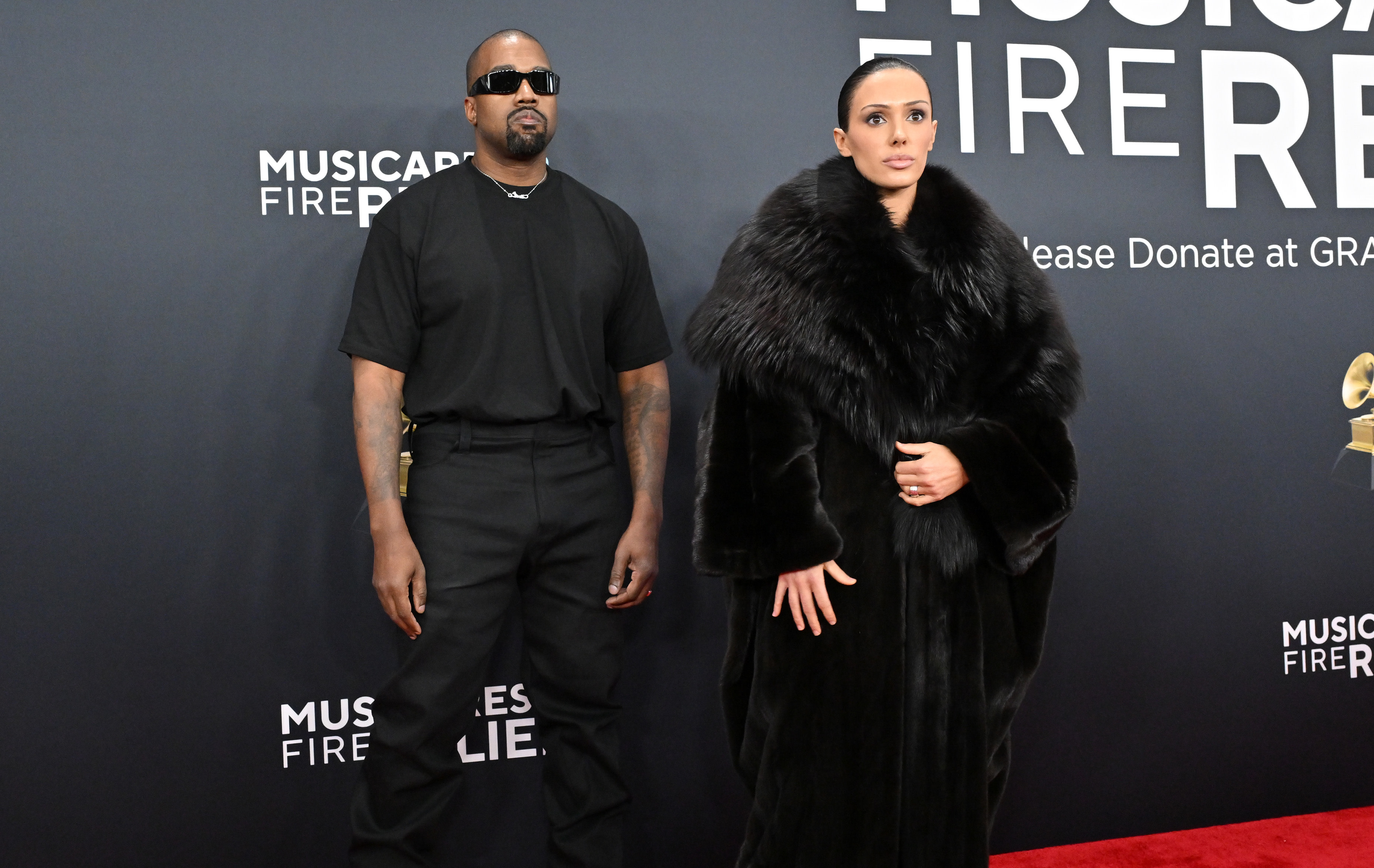 Kanye West wearing sunglasses and black attire, stands next to Bianca Censori in a black fur coat on a red carpet.