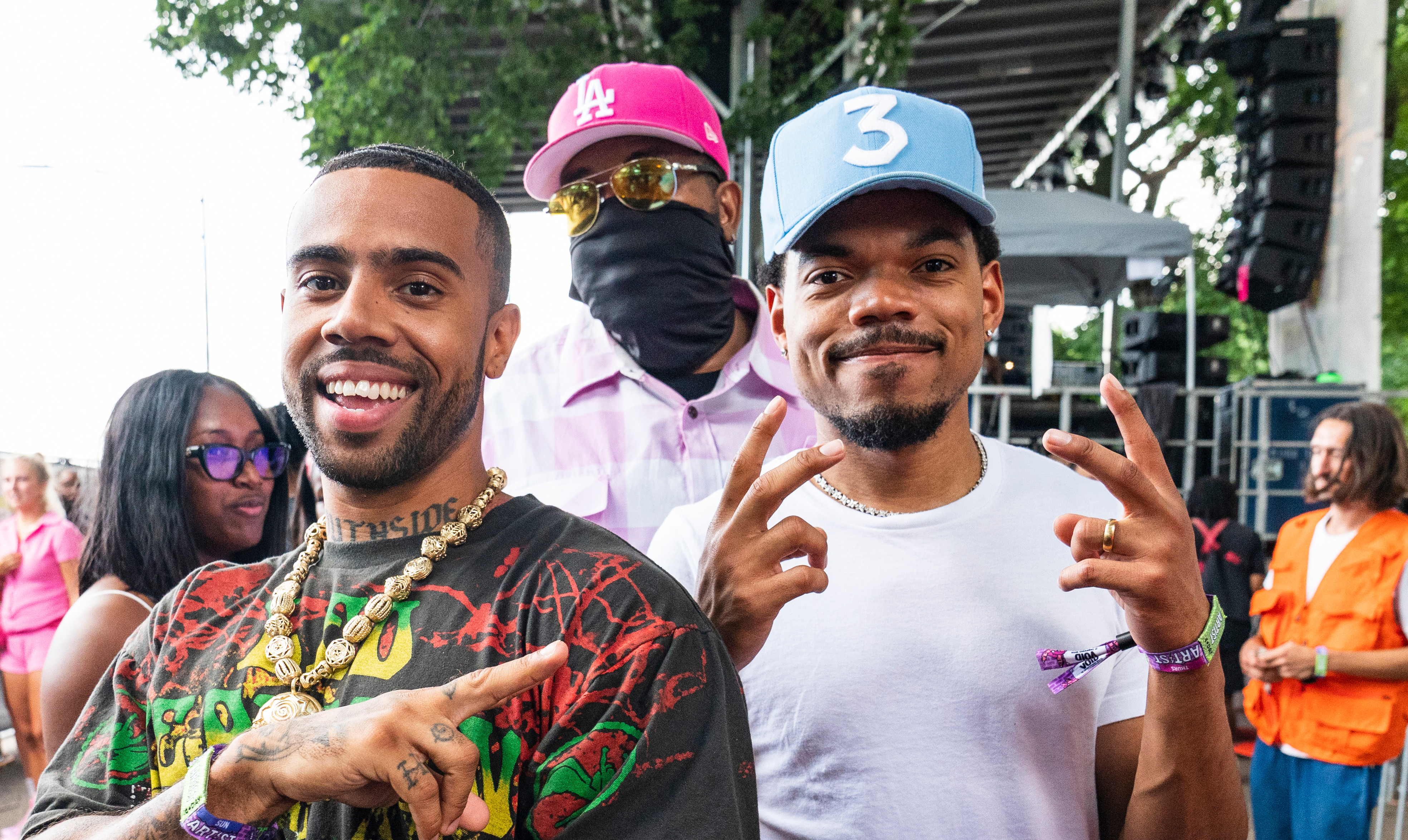 Vic Mensa and Chance the Rapper pose outdoors, smiling and making peace signs. A man in a mask and pink hat stands behind them.