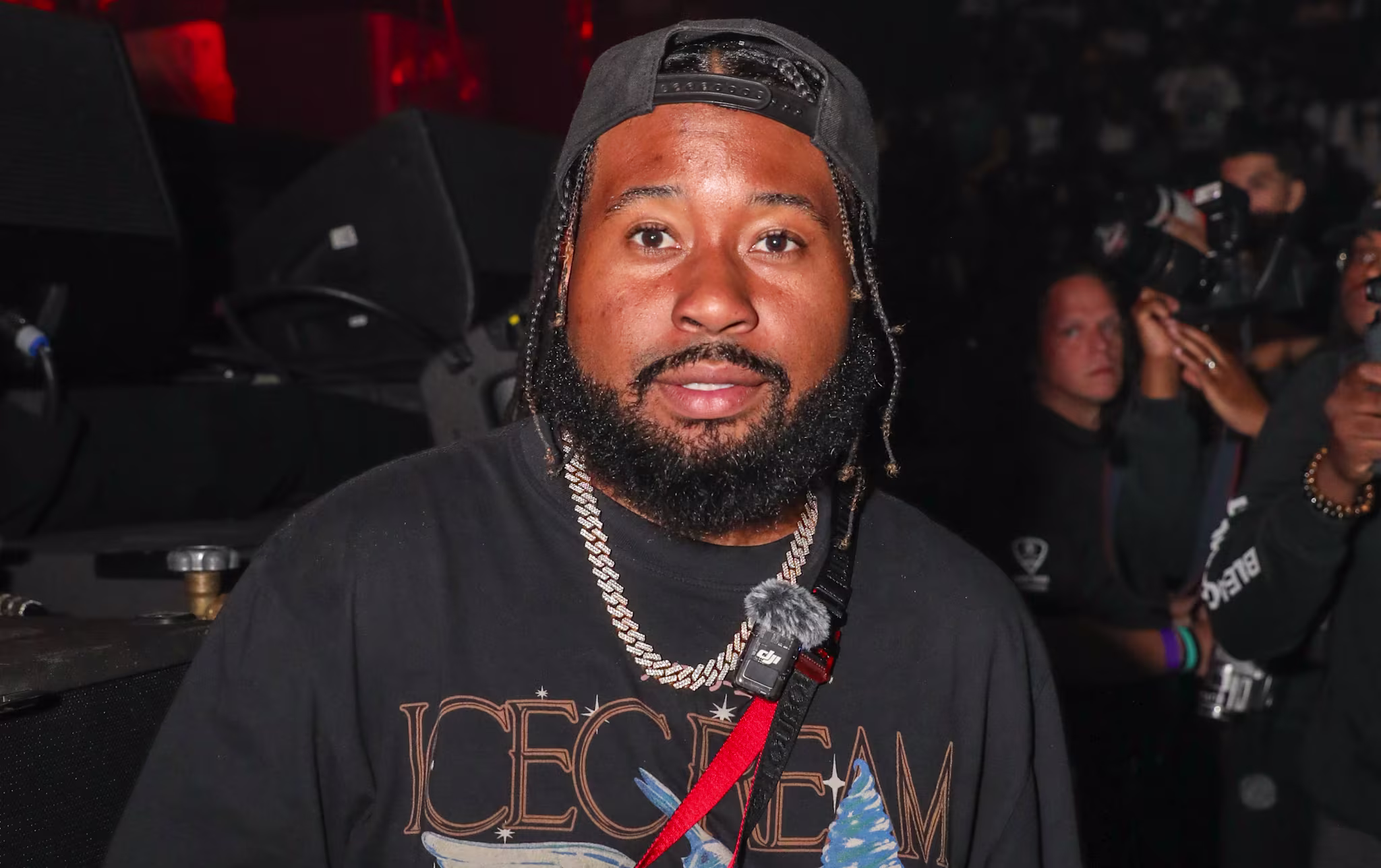 Akademiks wearing a black "Icecream" t-shirt and a cap, holding a phone, in a crowded event setting.