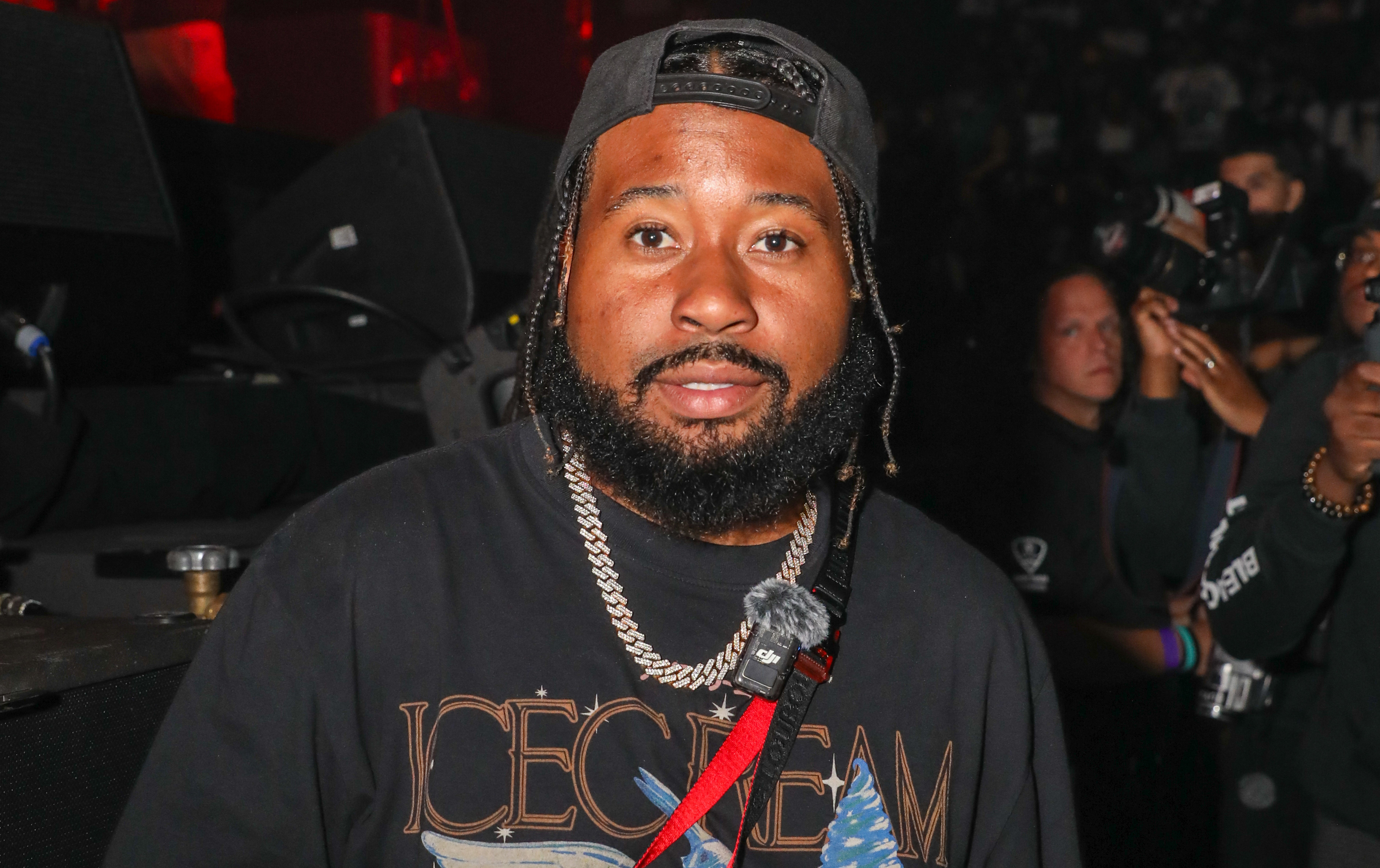 Akademiks wearing a black "Icecream" t-shirt and a cap, holding a phone, in a crowded event setting.