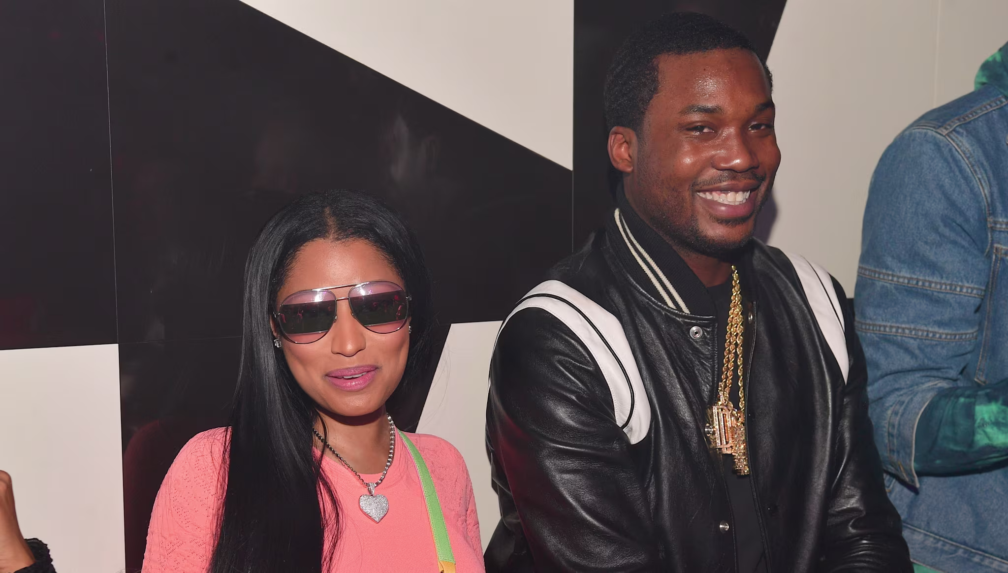 Nicki Minaj in sunglasses and pink outfit, sitting beside Meek Mill in a black jacket, both smiling at an event.