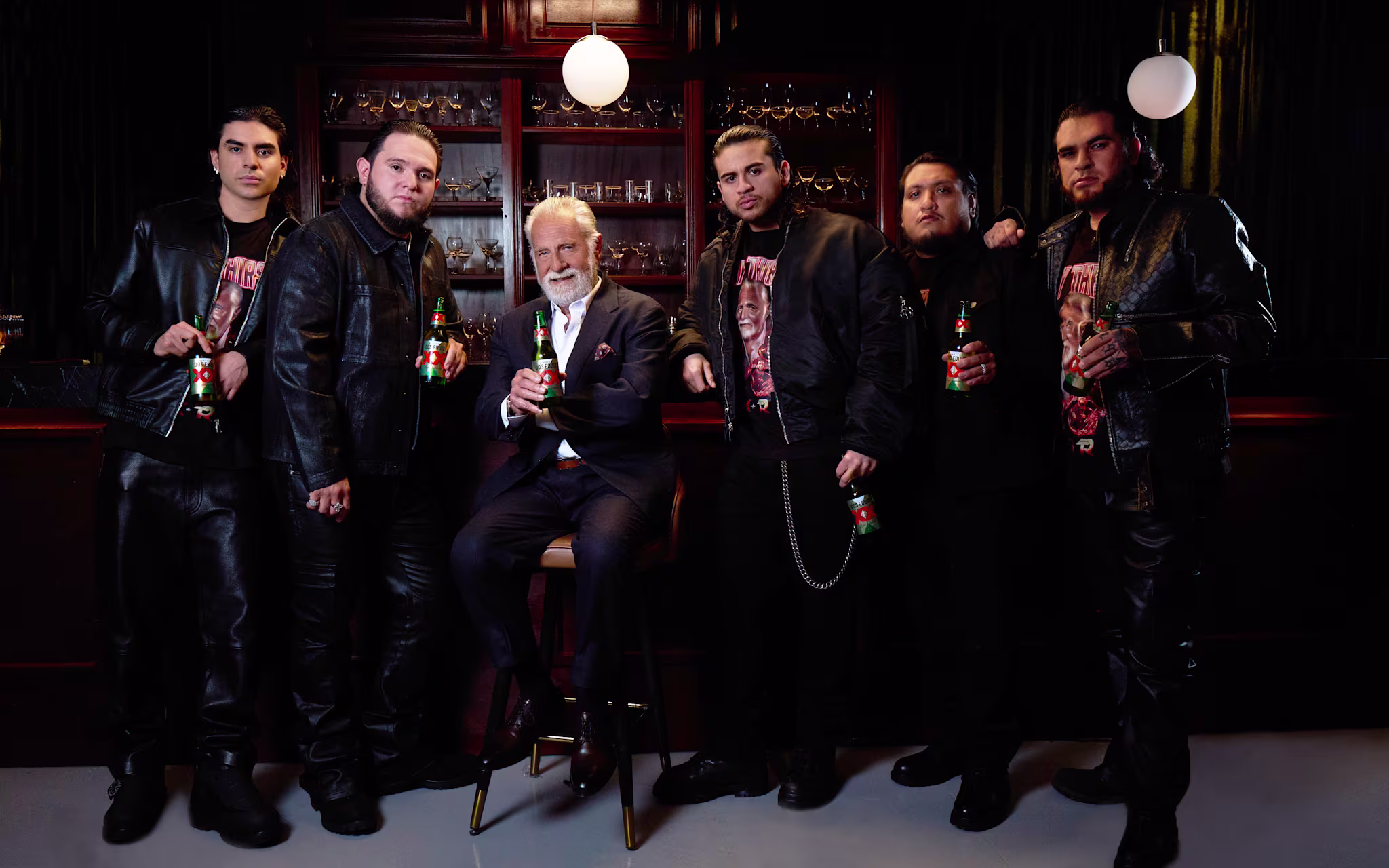 A group of men in leather jackets stand in a bar, holding drinks. A distinguished older man in a suit is seated among them.