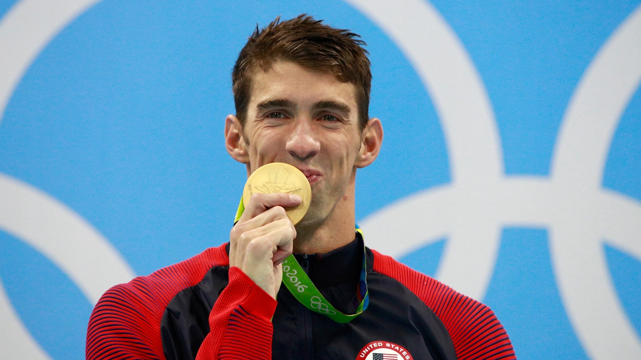 Michael Phelps, wearing a Team USA jacket, bites a gold medal at the 2016 Olympics, with Olympic rings in the background.