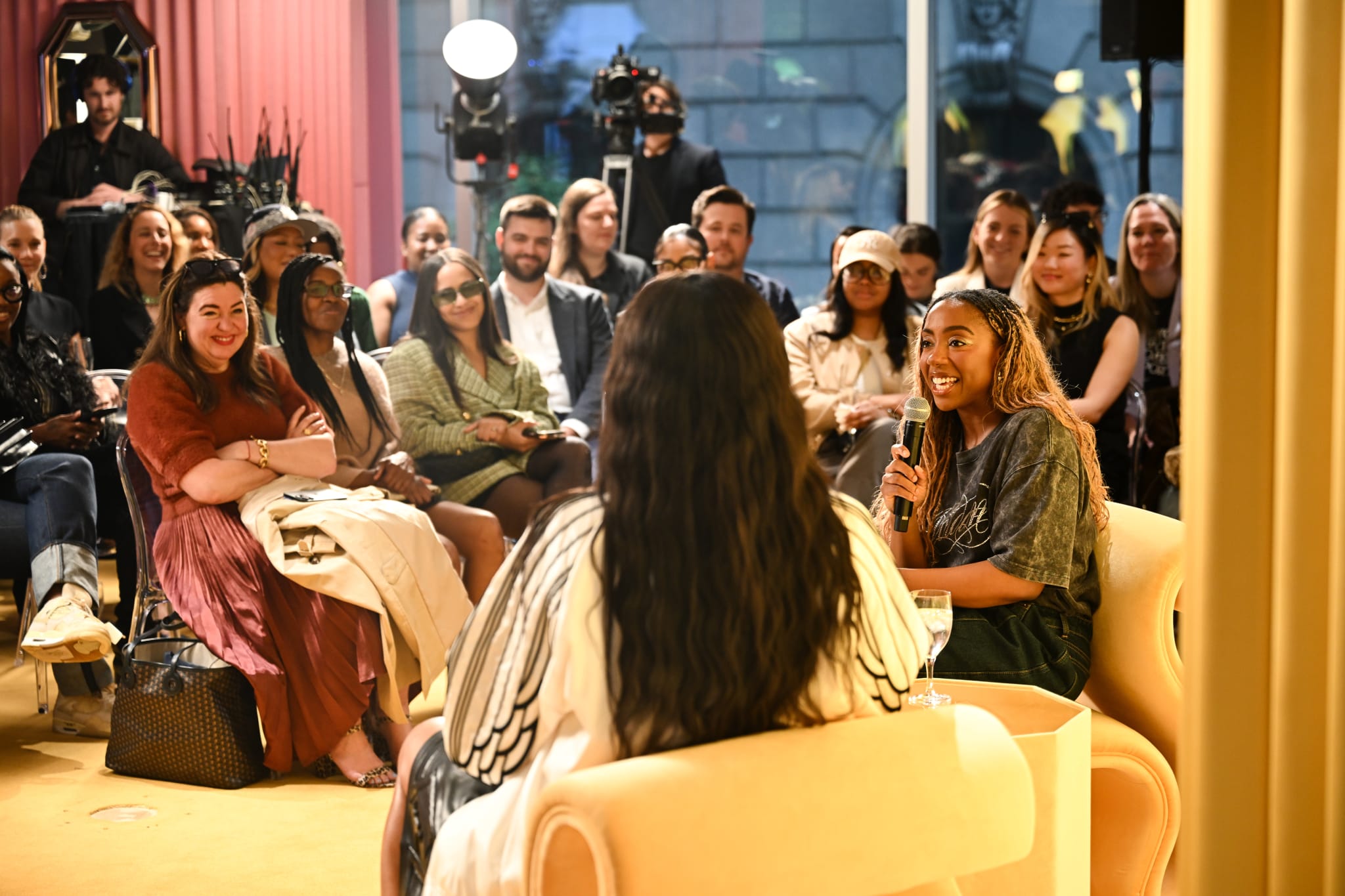 A woman speaks into a microphone in a cozy room with an audience seated around, engaged and listening attentively.