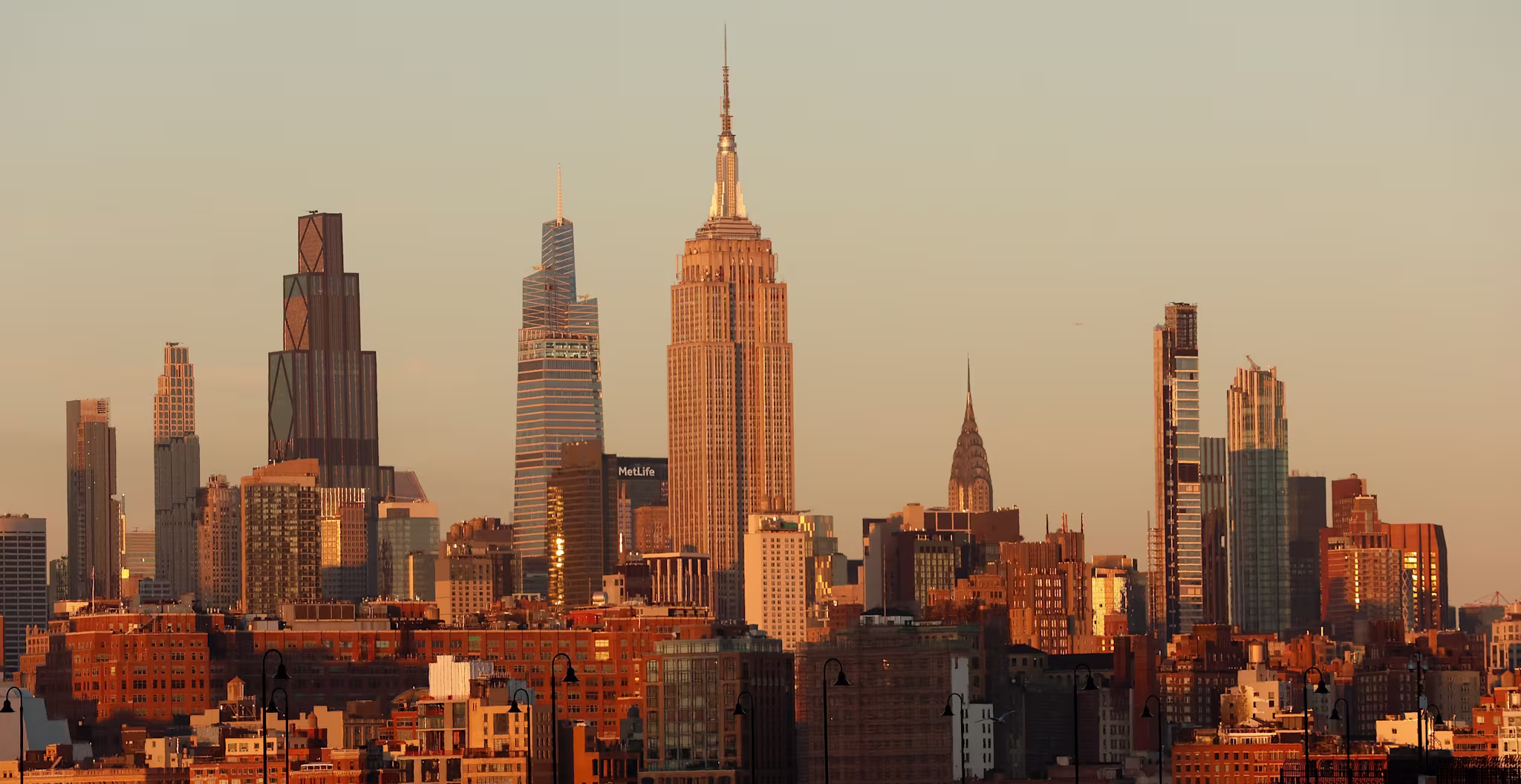 New York City skyline at sunset, featuring the Empire State Building and surrounding skyscrapers bathed in golden light.