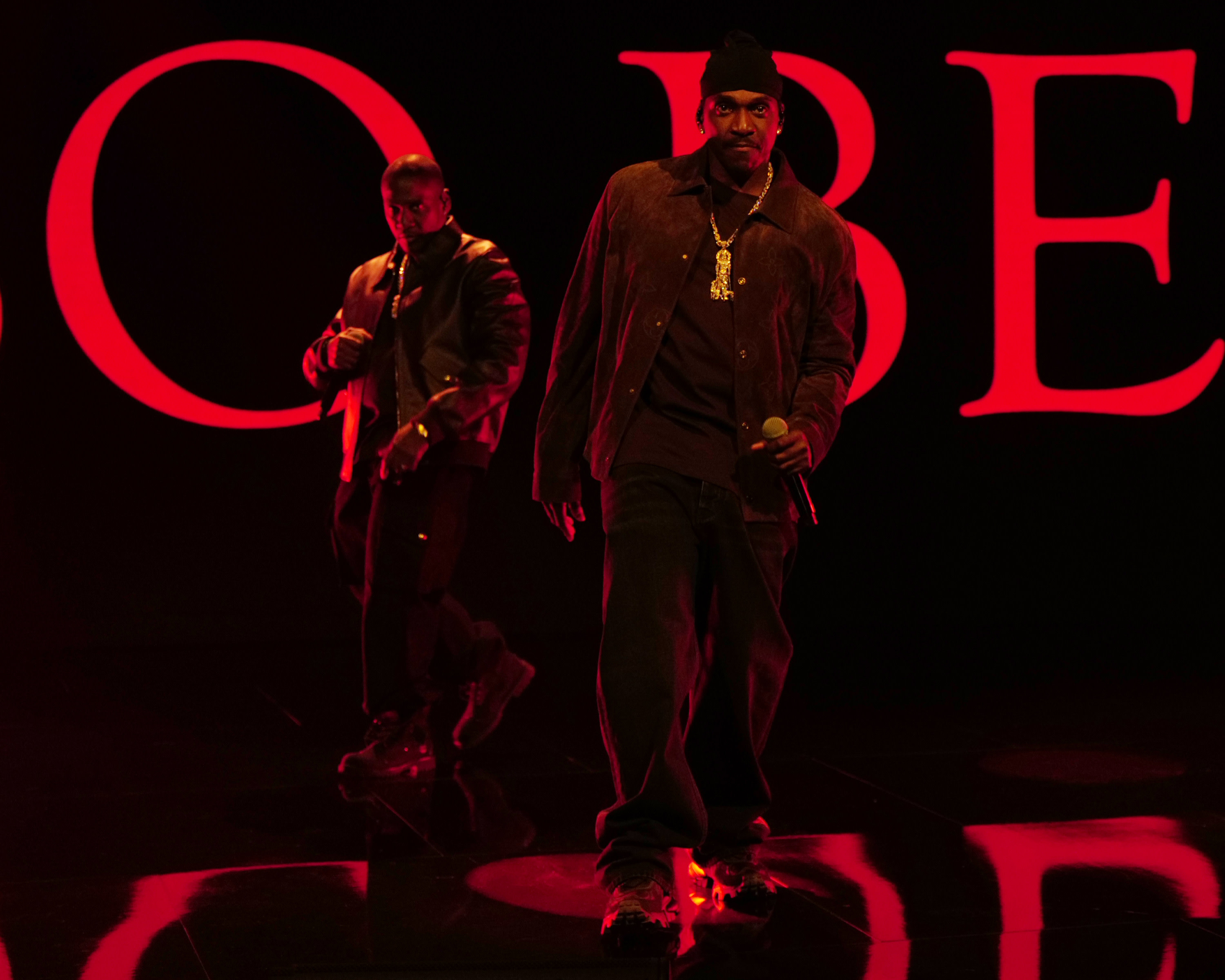 Clipse on stage with red lighting, holding microphones, wearing dark clothing and beanies. Large red letters in the background.