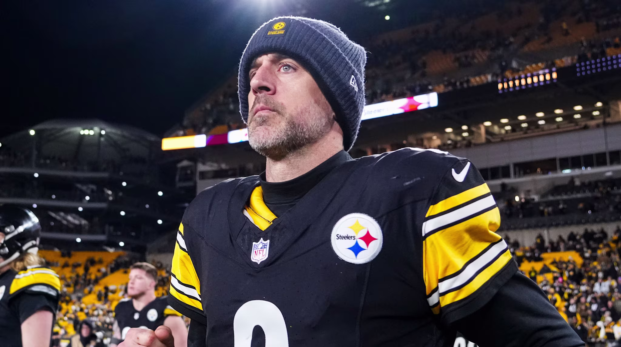 Aaron Rodgers in Pittsburgh Steelers uniform on the field with a stadium full of fans in the background.