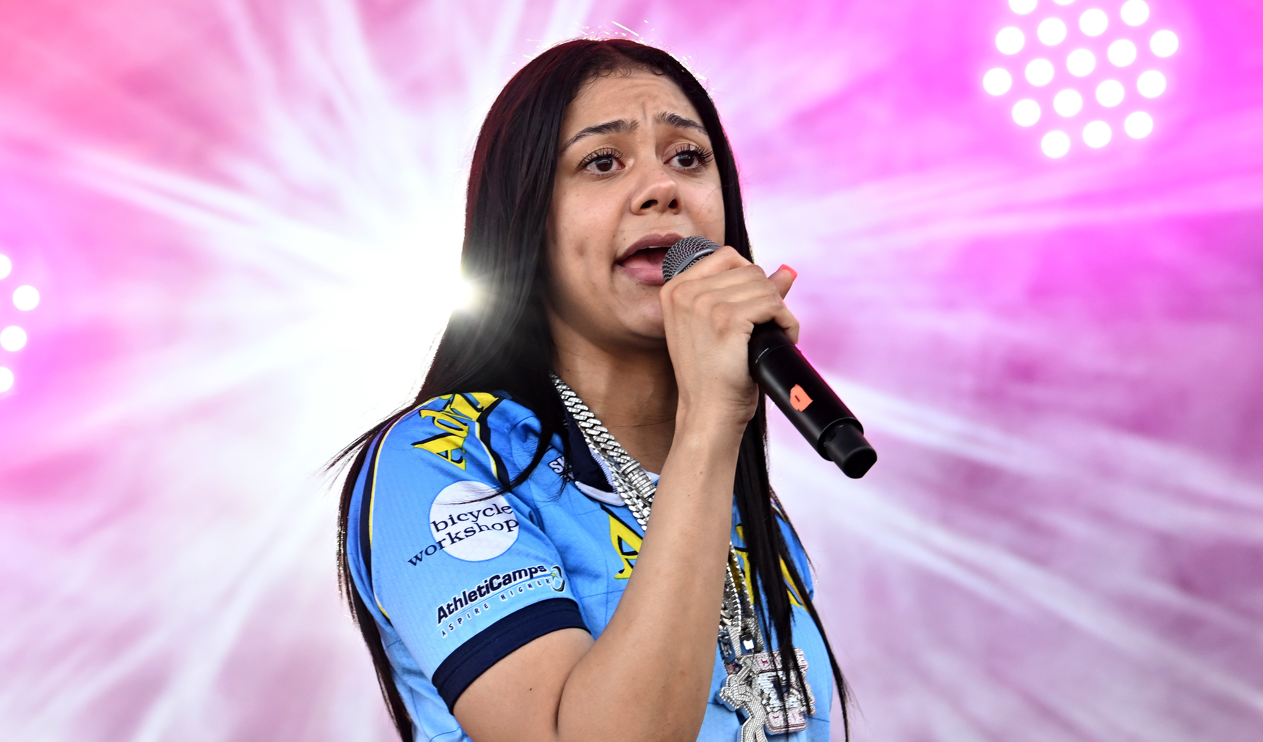 Karrahbooo singing on stage with a microphone, wearing a blue and white shirt, against a backdrop of pink and white lights.