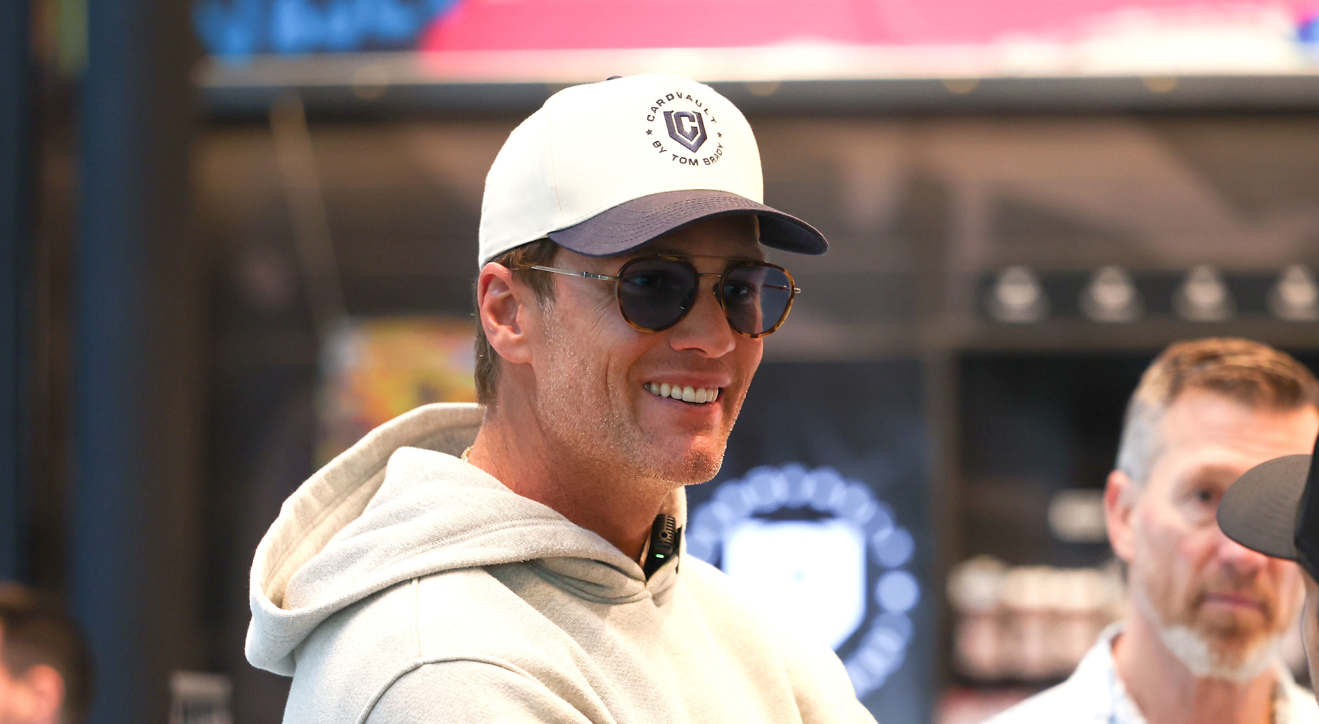 A man in a white cap and sunglasses smiles while talking to others indoors.