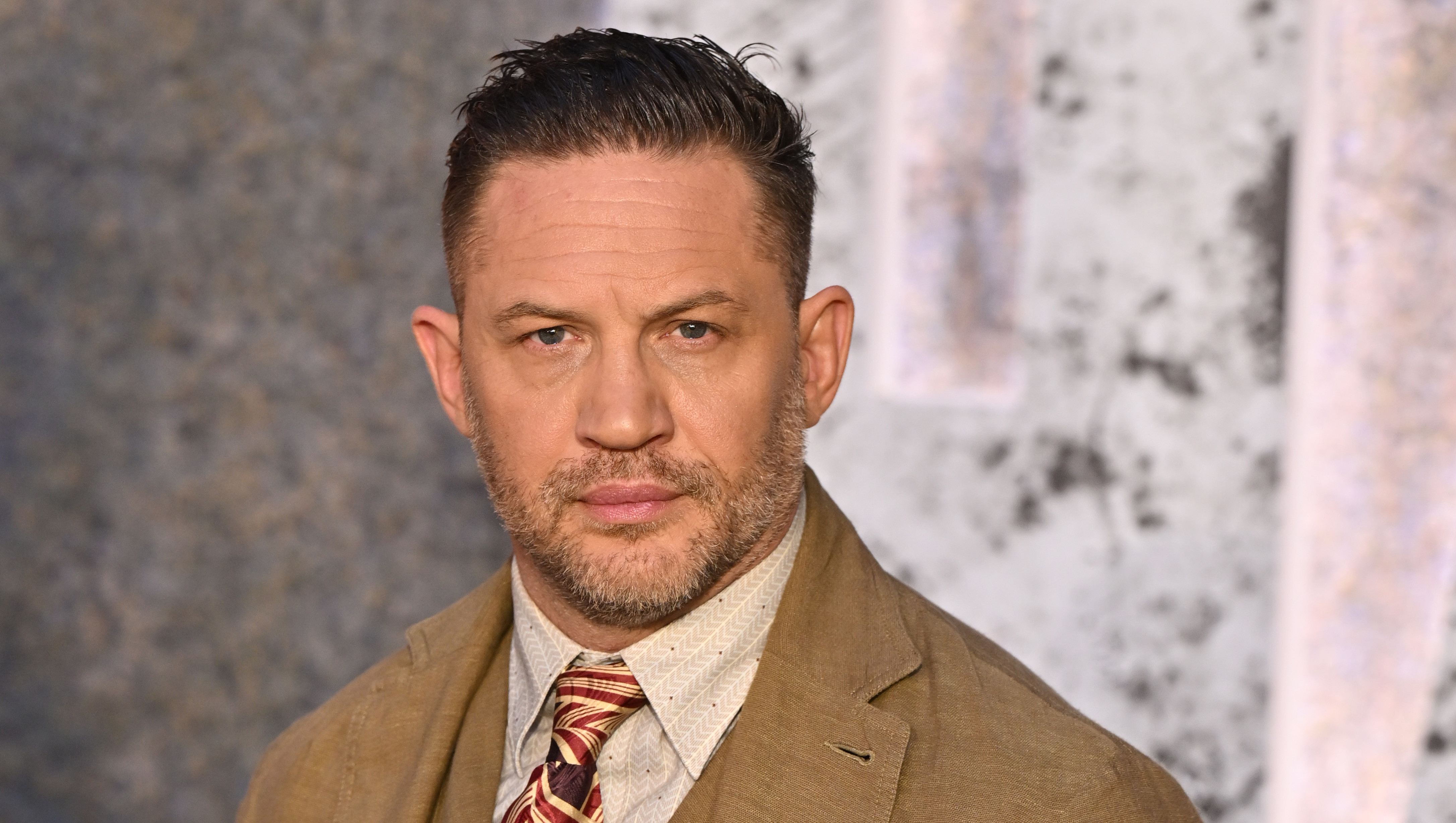 Tom Hardy in a brown suit with a patterned tie, posing with hands in pockets against a textured backdrop.