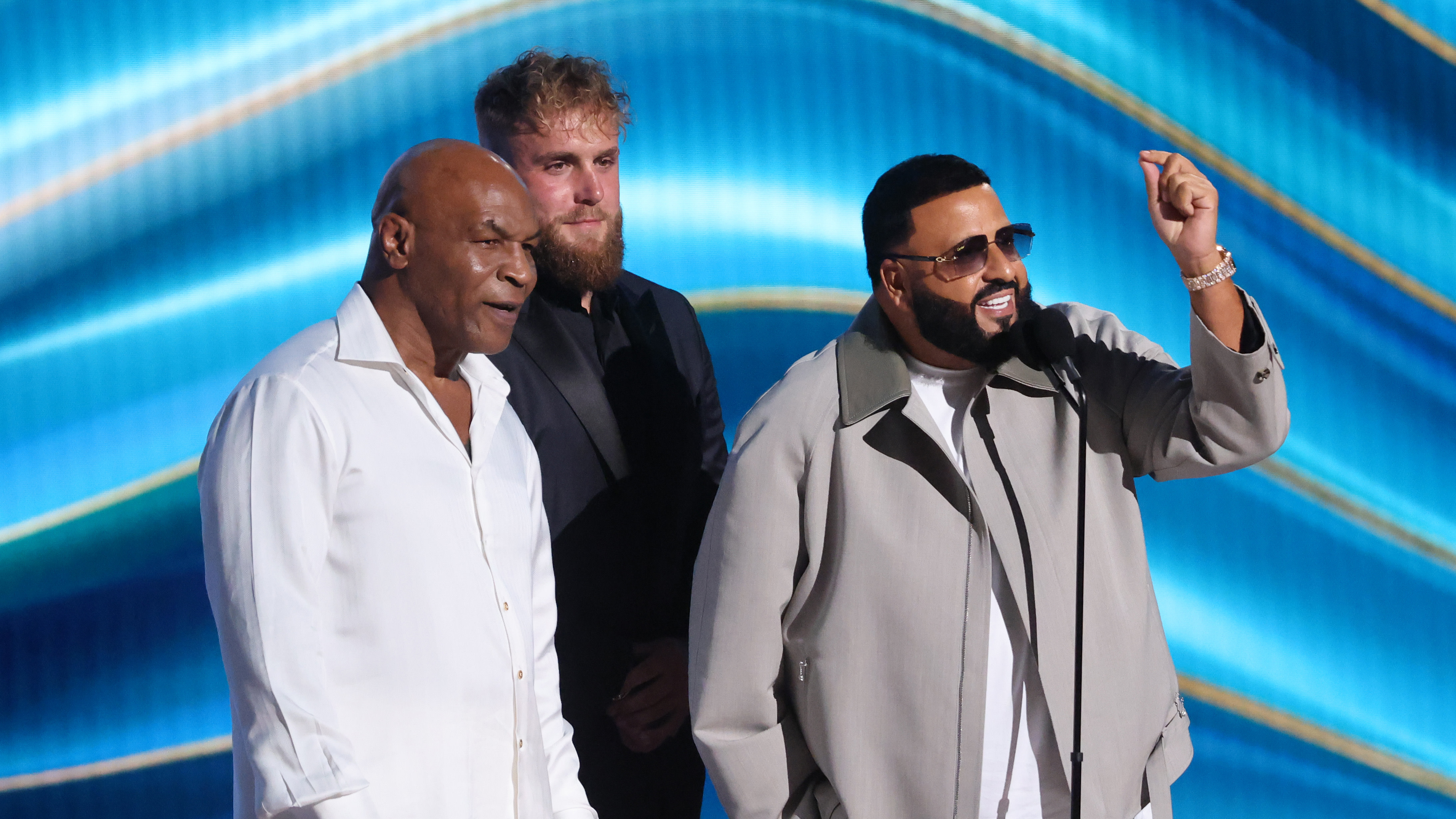 Mike Tyson, Jake Paul, and DJ Khaled on stage, smiling and interacting with the audience against a blue backdrop.