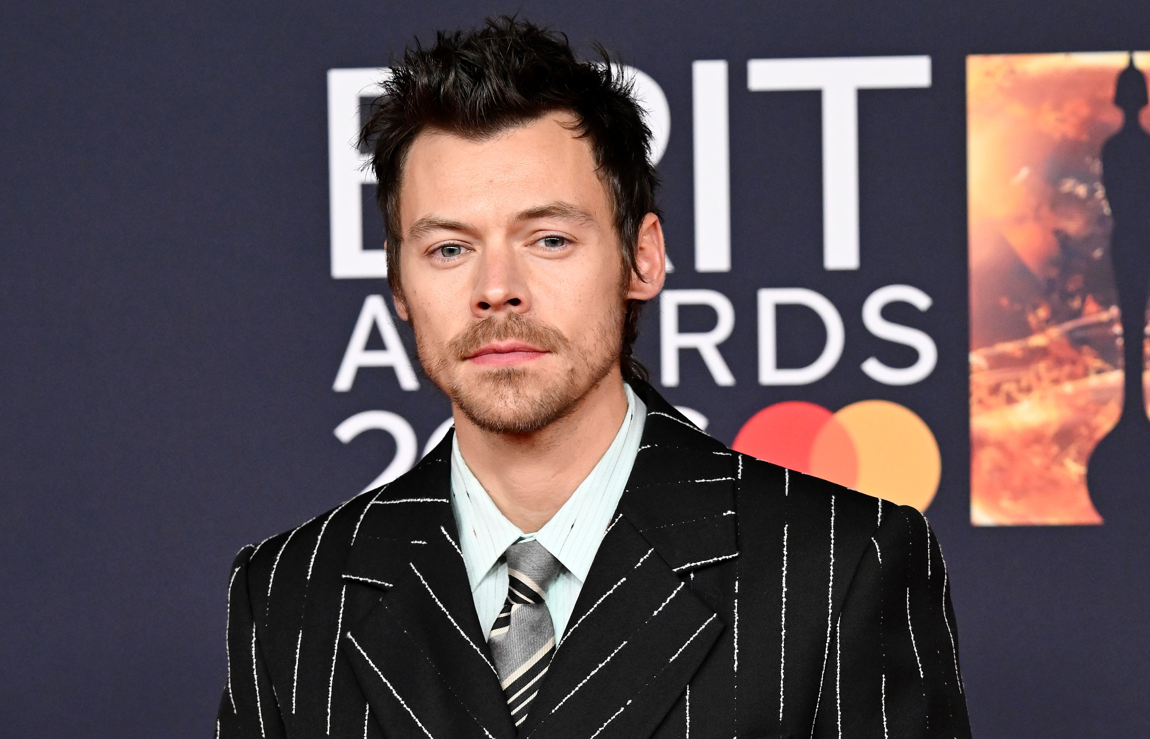 Harry Styles at the BRIT Awards, wearing a black pinstripe suit with a striped tie, standing against a branded backdrop.