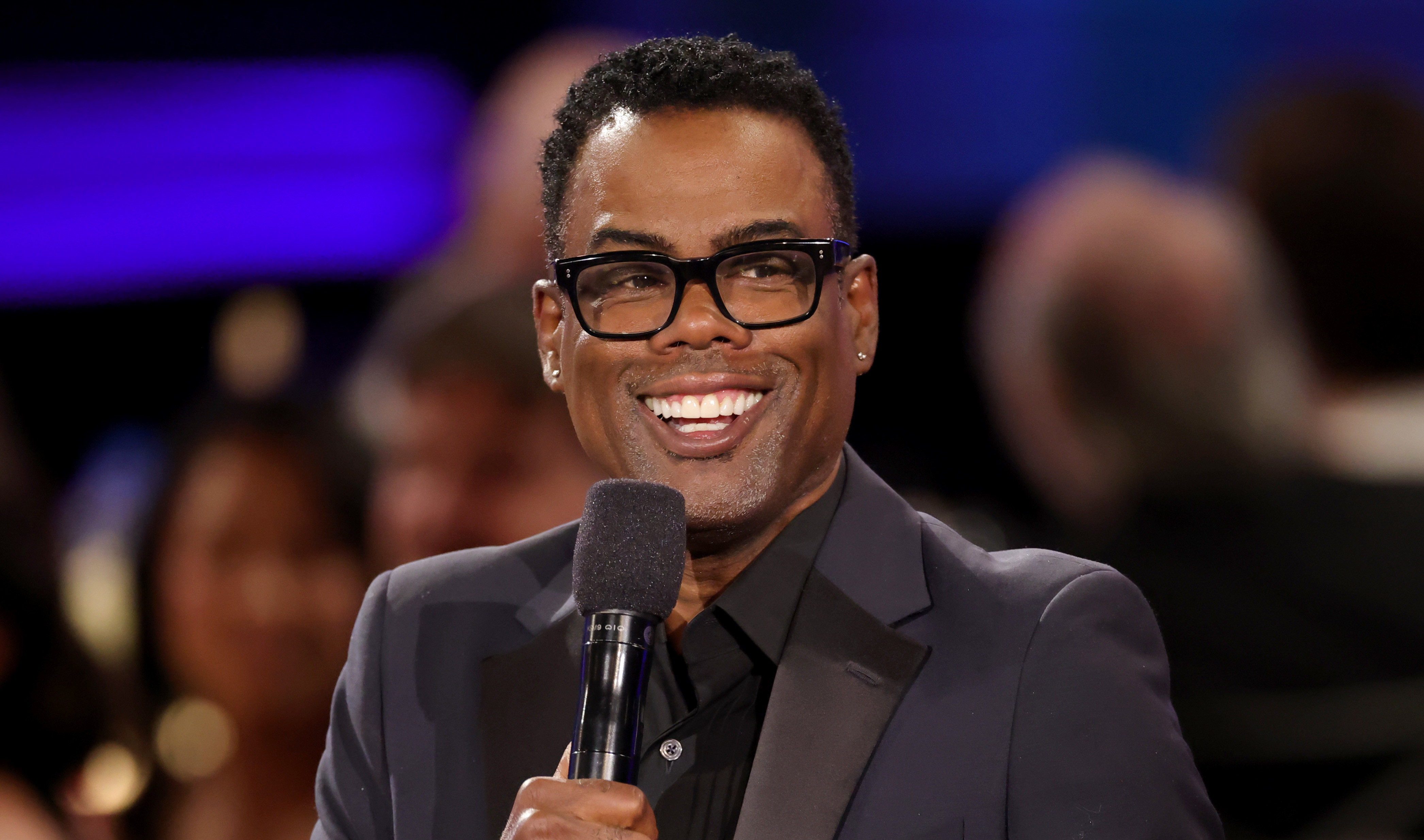 Chris Rock, wearing glasses and a dark suit, smiling and holding a microphone at an event.