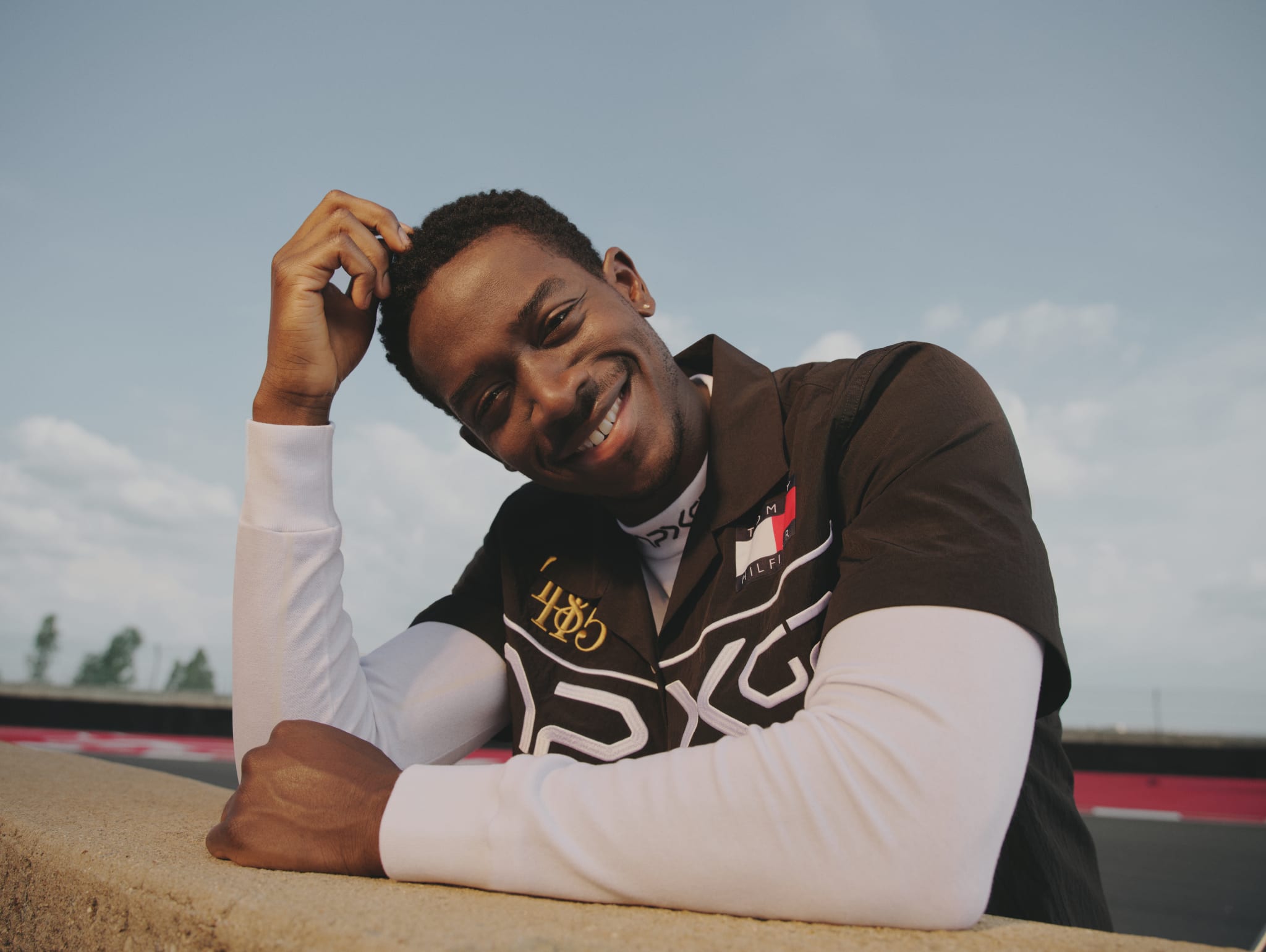 A person smiling, leaning on a ledge outdoors, wearing a black and white shirt with logos. The sky is clear with some clouds.