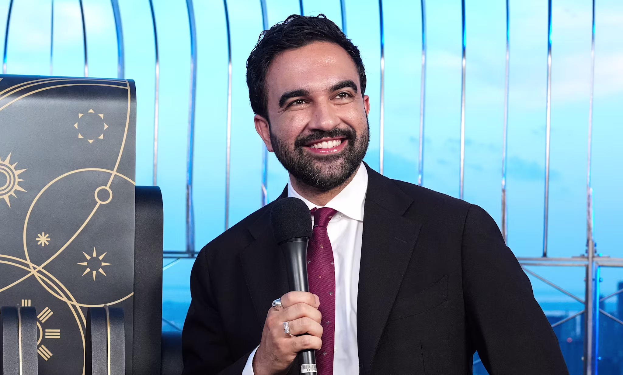 Zohran Mamdani in a suit holds a microphone, smiling, with a city skyline in the background.