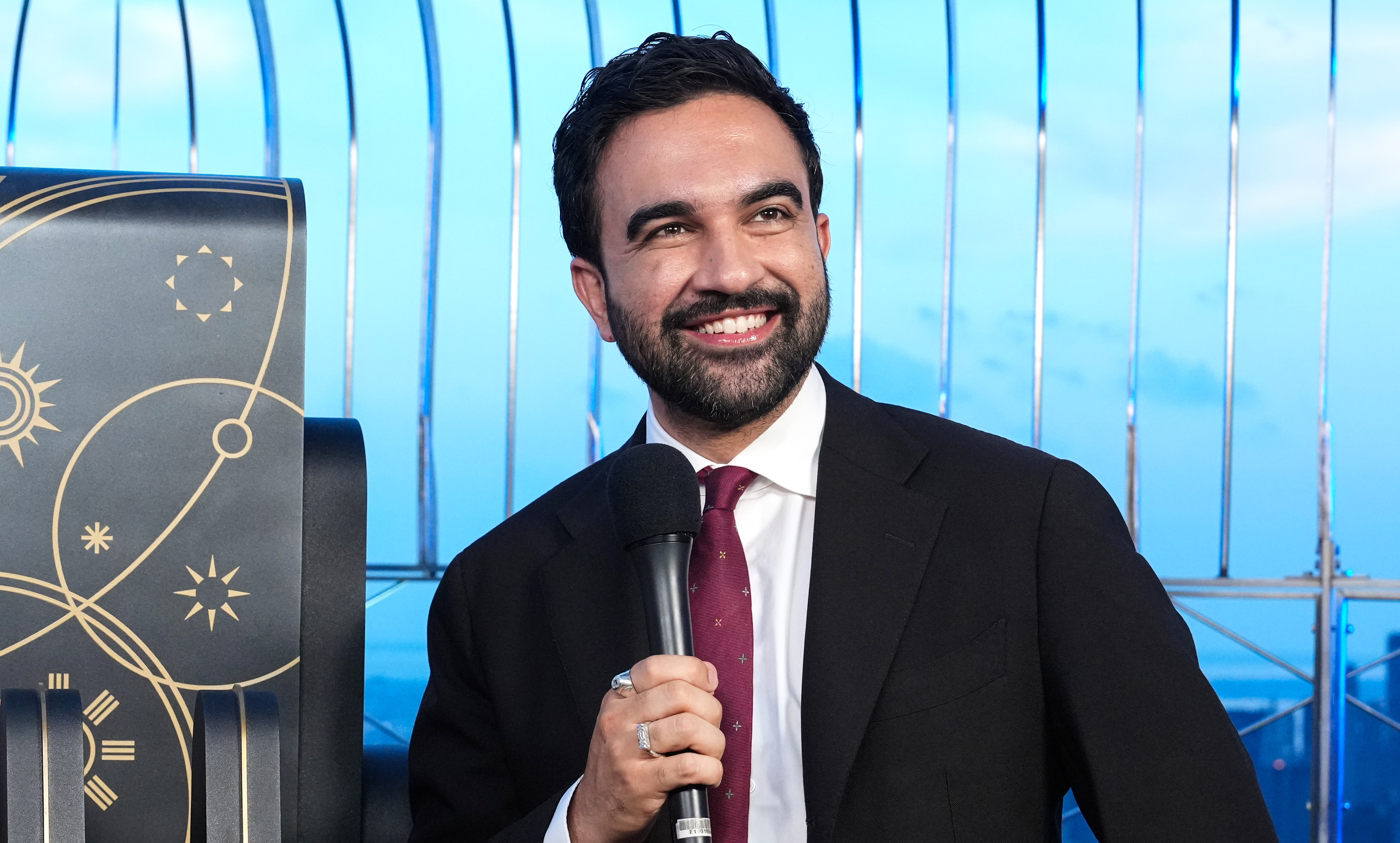 Zohran Mamdani in a suit holds a microphone, smiling, with a city skyline in the background.