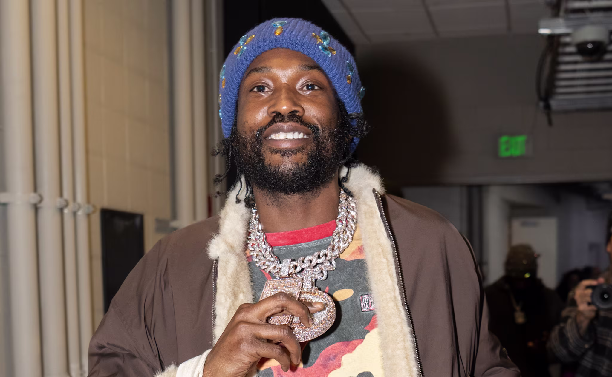 Meek Mill wearing a colorful beanie, large chain necklace, and a camouflage shirt holds a drink. He is in a hallway with people in the background.