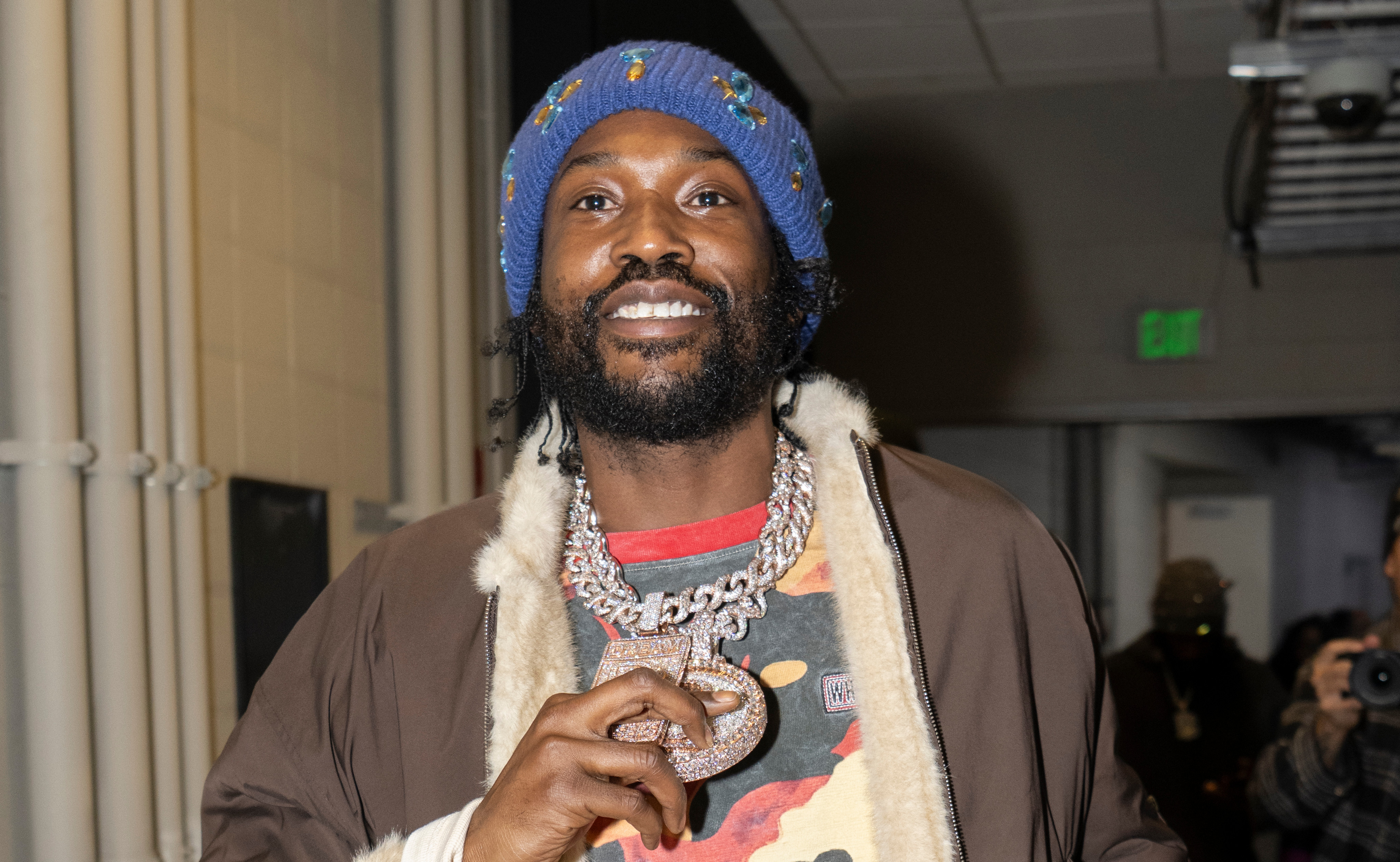 Meek Mill wearing a colorful beanie, fur-lined jacket, and camouflage shirt holds a drink and a large chain in a hallway.
