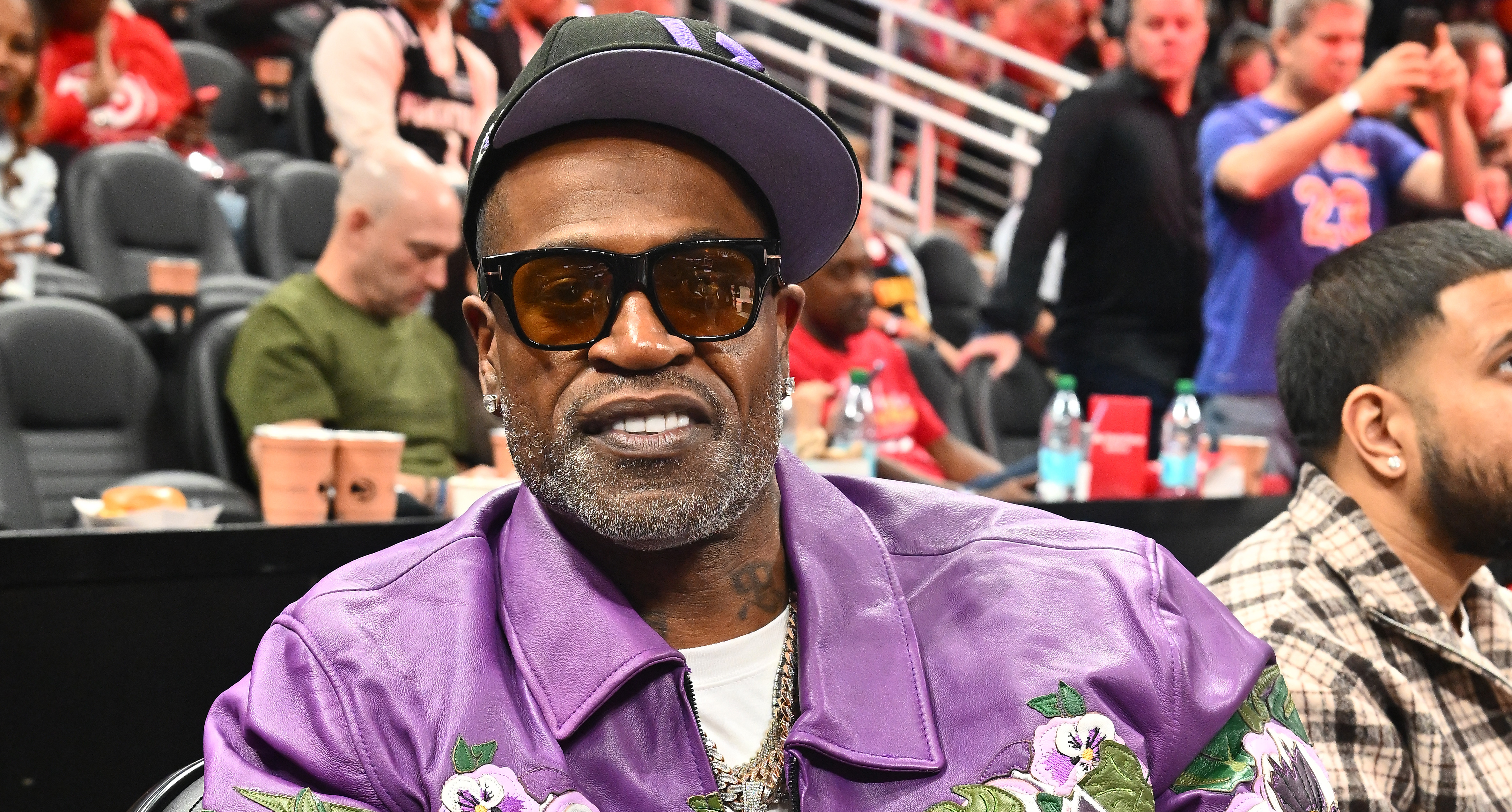 Stephen Jackson in a purple floral jacket and sunglasses sits courtside at a basketball game, flashing a peace sign.