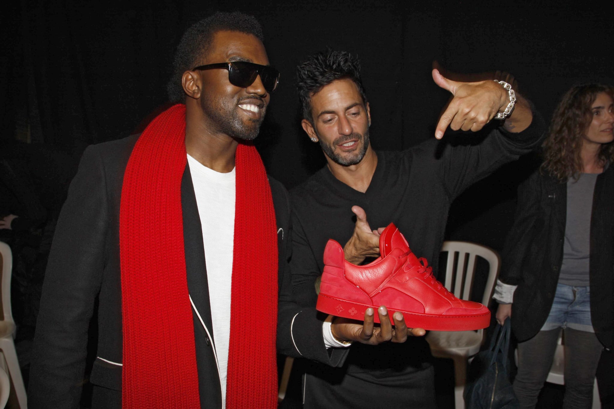 Kanye West, wearing sunglasses and a red scarf, holds a red Louis Vuitton Don with designer Marc Jacobs pointing at it.