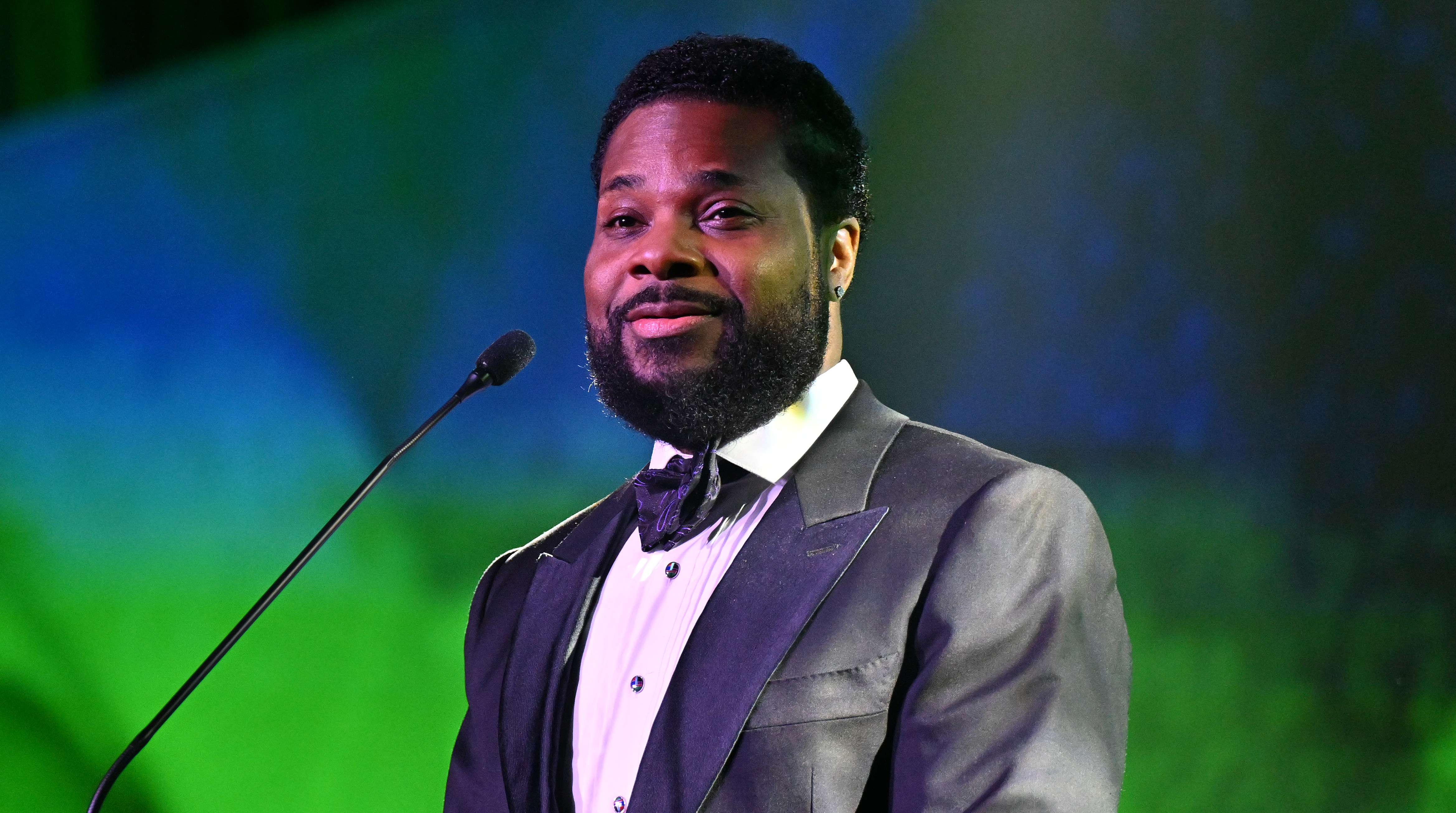 Malcolm-Jamal Warner in a suit and bow tie speaks at a podium with a green backdrop.