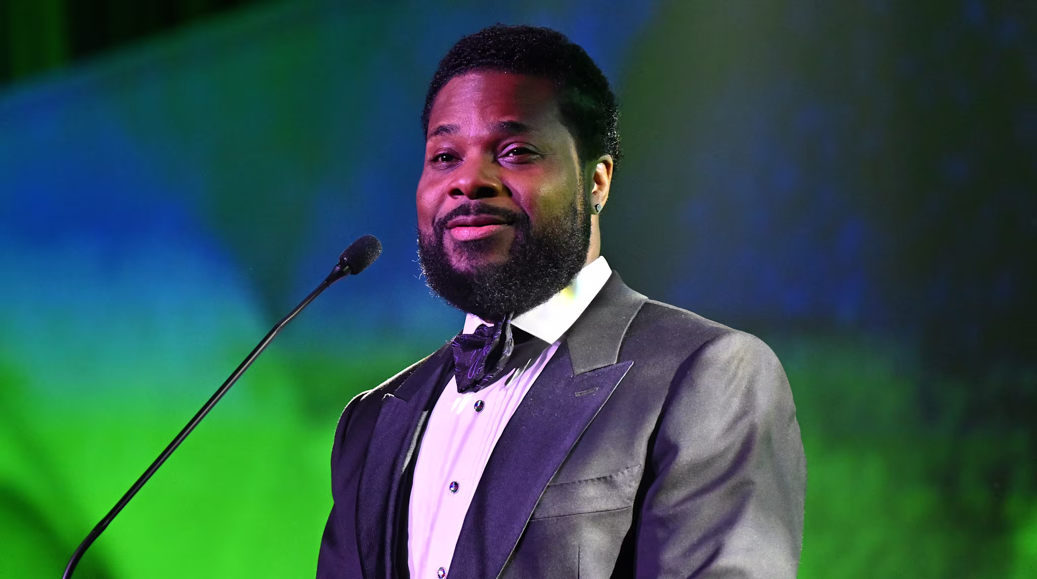 Malcolm-Jamal Warner in a suit and bow tie speaks at a podium with a green backdrop.