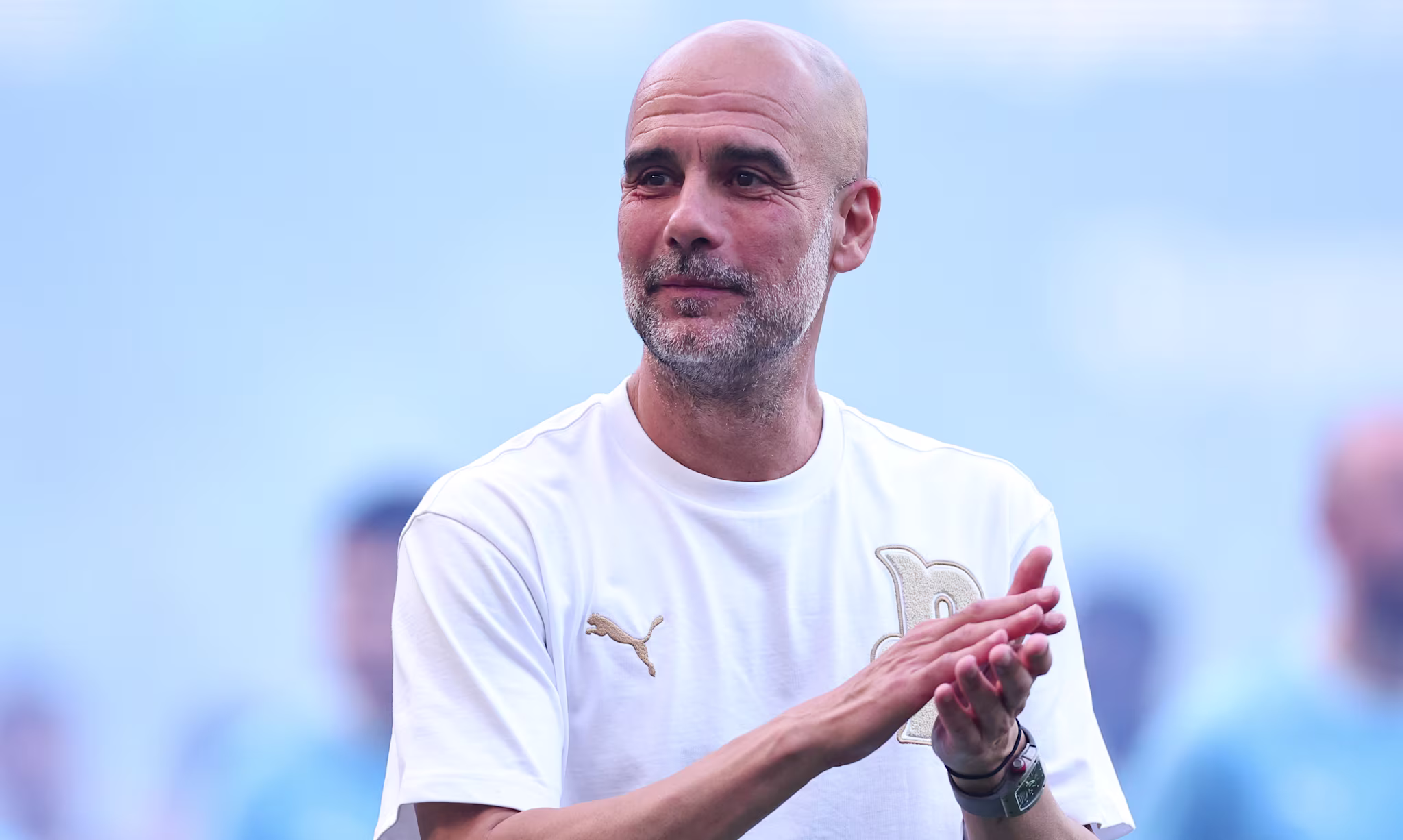 Pep Guardiola in a white Puma shirt clapping, with a blurred blue background.