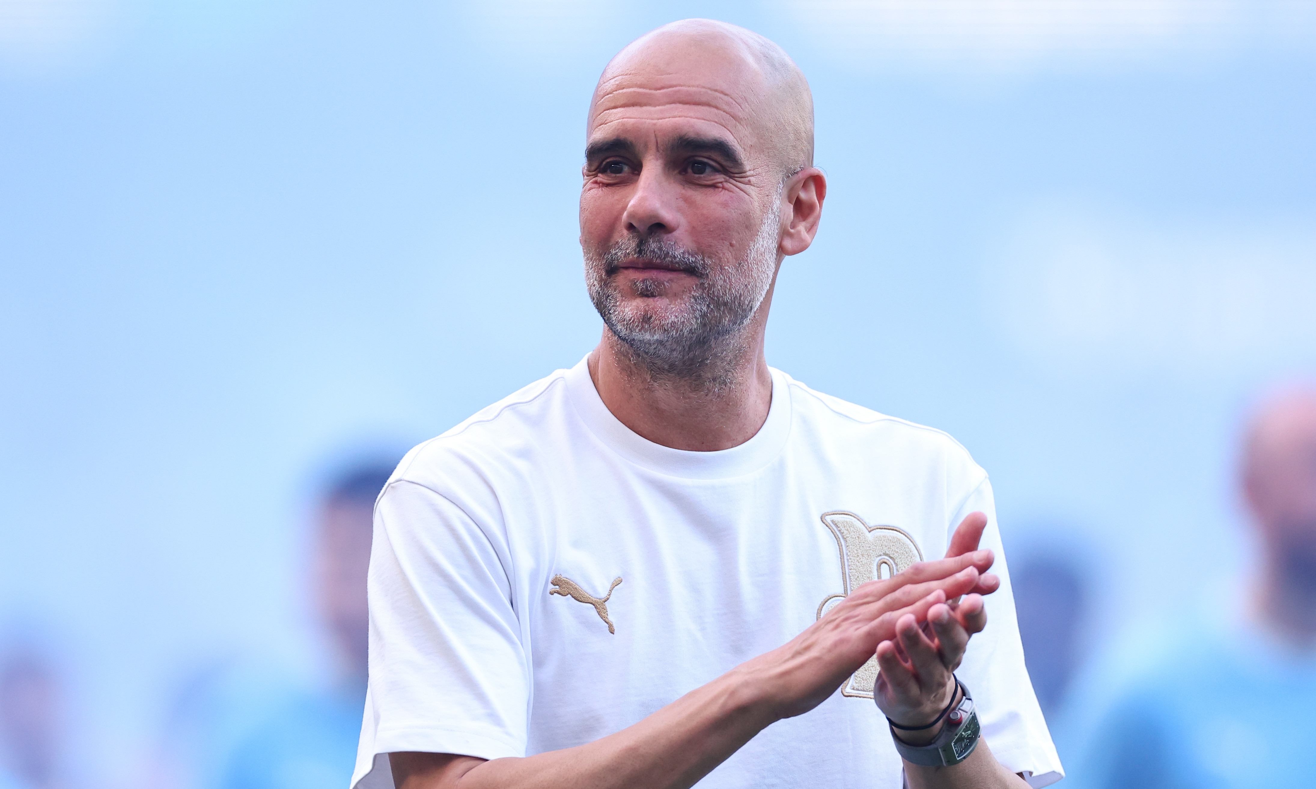 Pep Guardiola in a white Puma shirt clapping, with a blurred blue background.