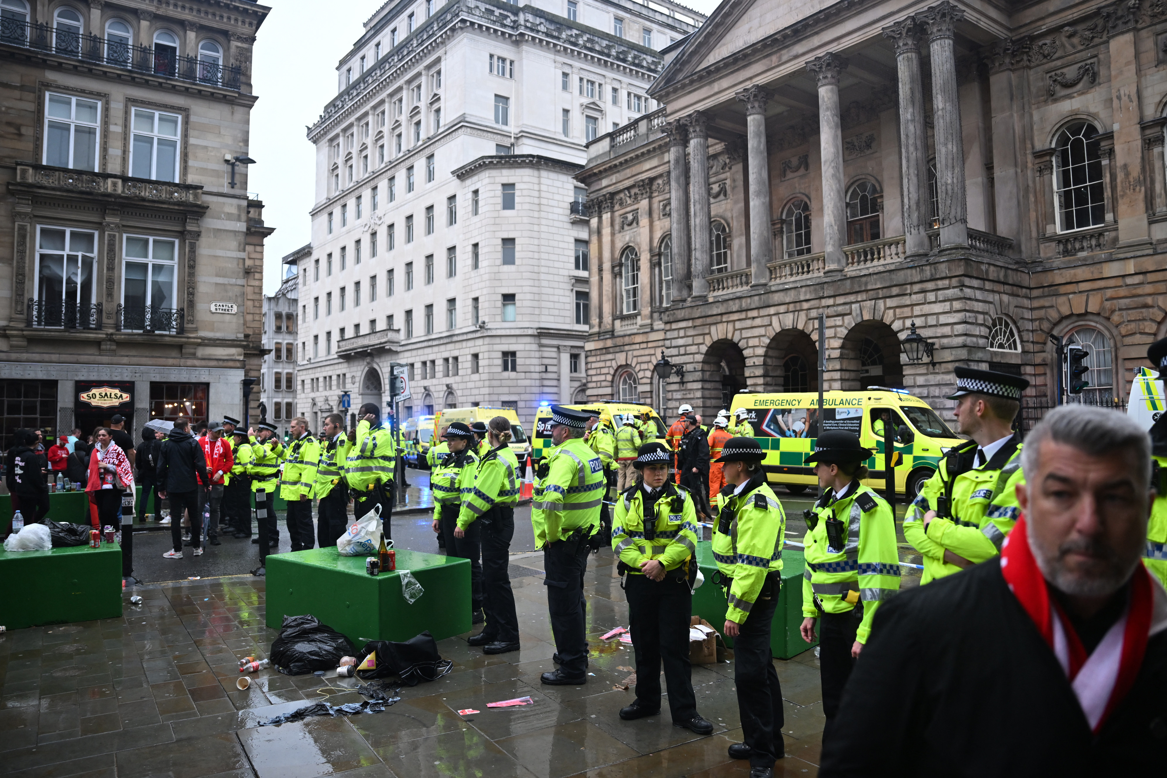 Police officers in high-visibility jackets stand in a city square with historic buildings. Ambulances are parked nearby, and people are gathered around.