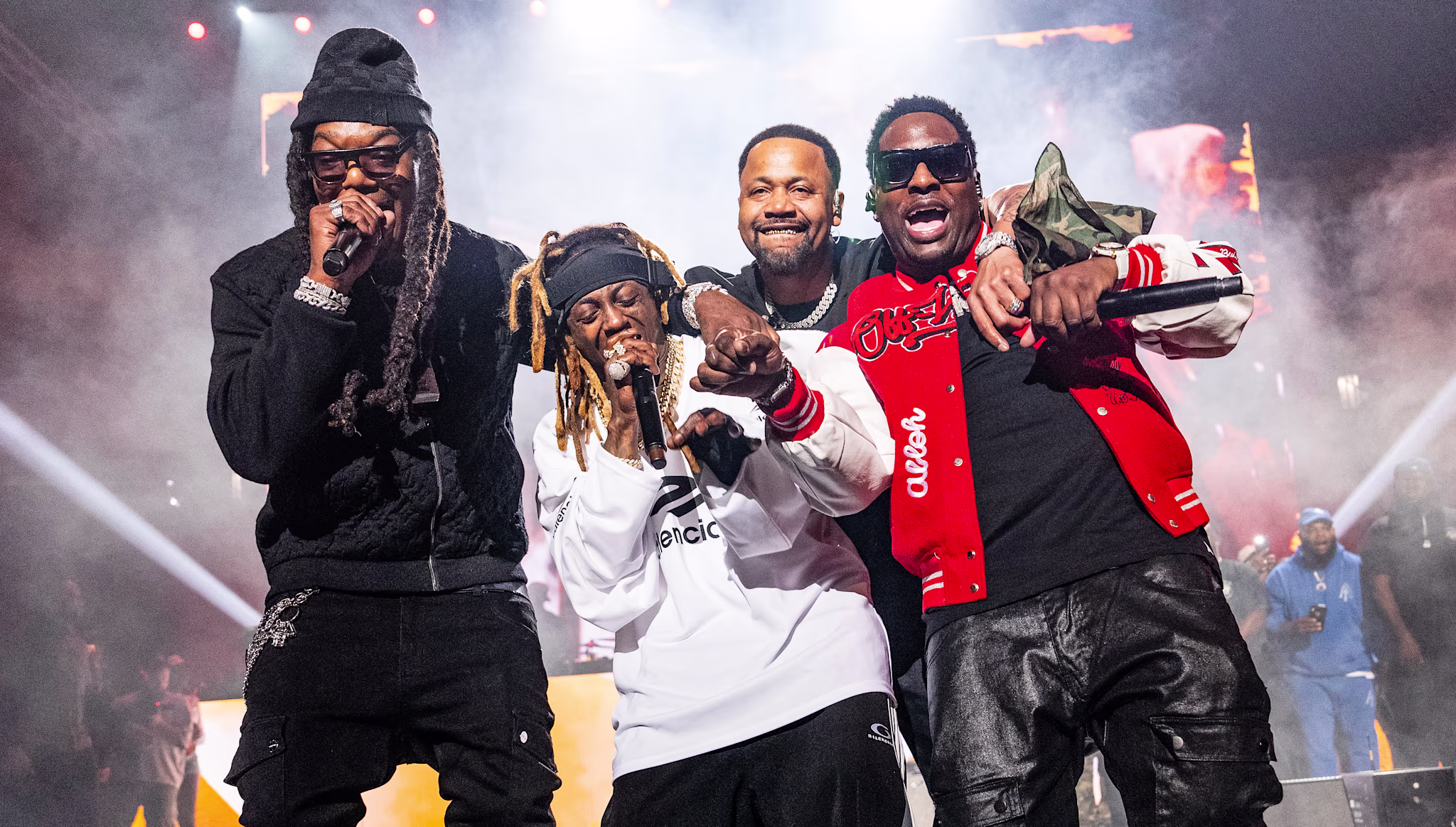 BG, Turk, Juvenile and Lil Wayne in a white sweatshirt, performing energetically with microphones against a backdrop of lights and smoke.