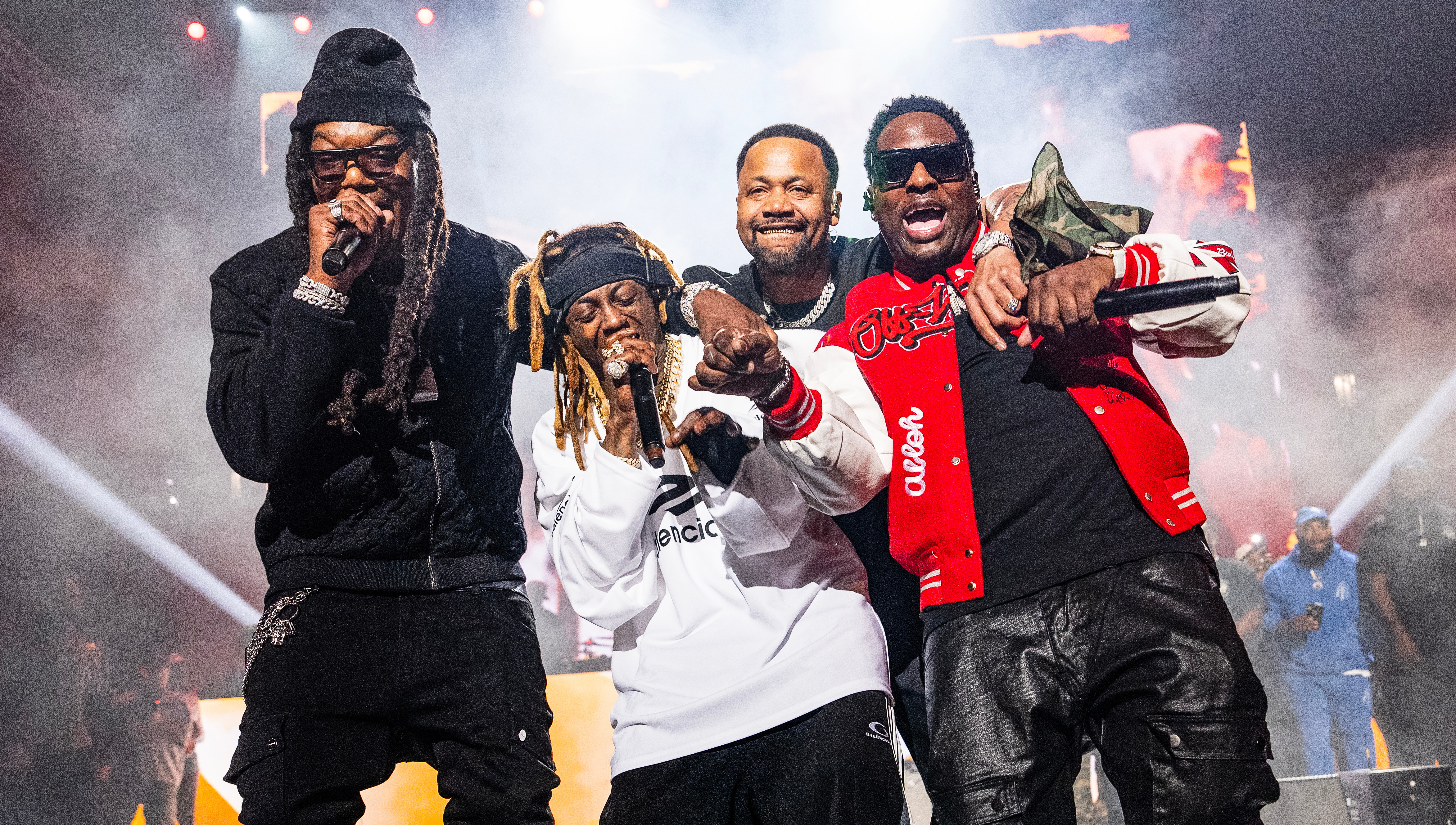 BG, Turk, Juvenile and Lil Wayne in a white sweatshirt, performing energetically with microphones against a backdrop of lights and smoke.