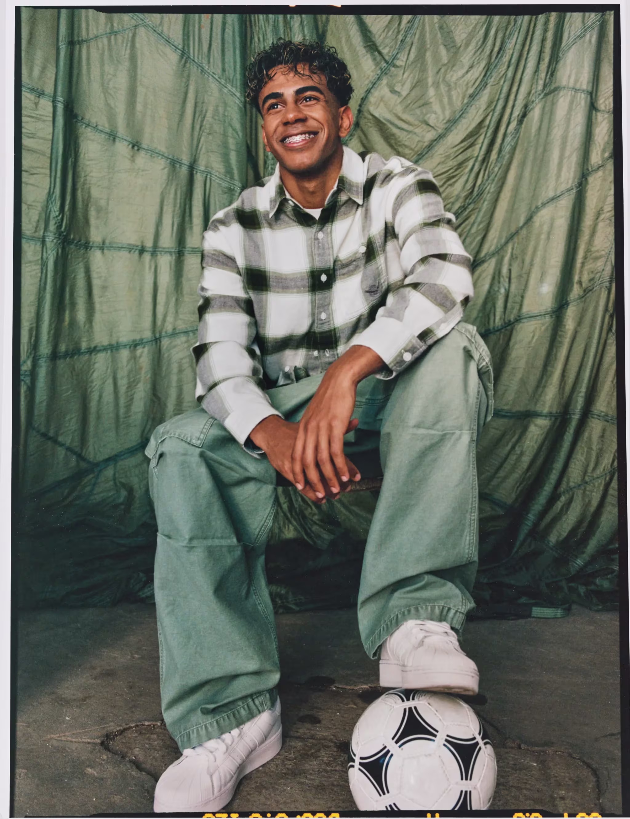 A person in a plaid shirt and green pants sits smiling with a soccer ball at their feet, against a fabric backdrop.