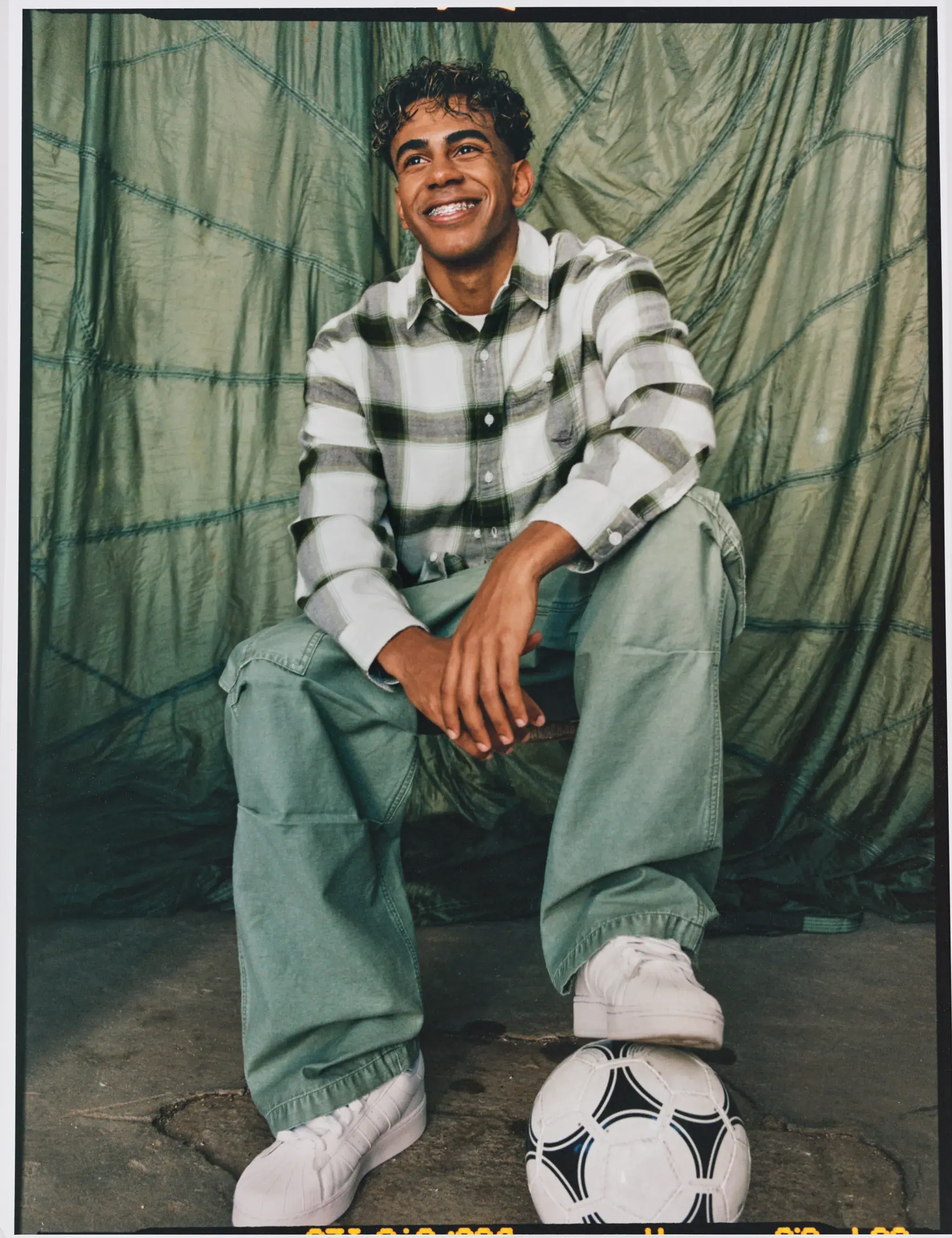 A person in a plaid shirt and green pants sits smiling with a soccer ball at their feet, against a fabric backdrop.
