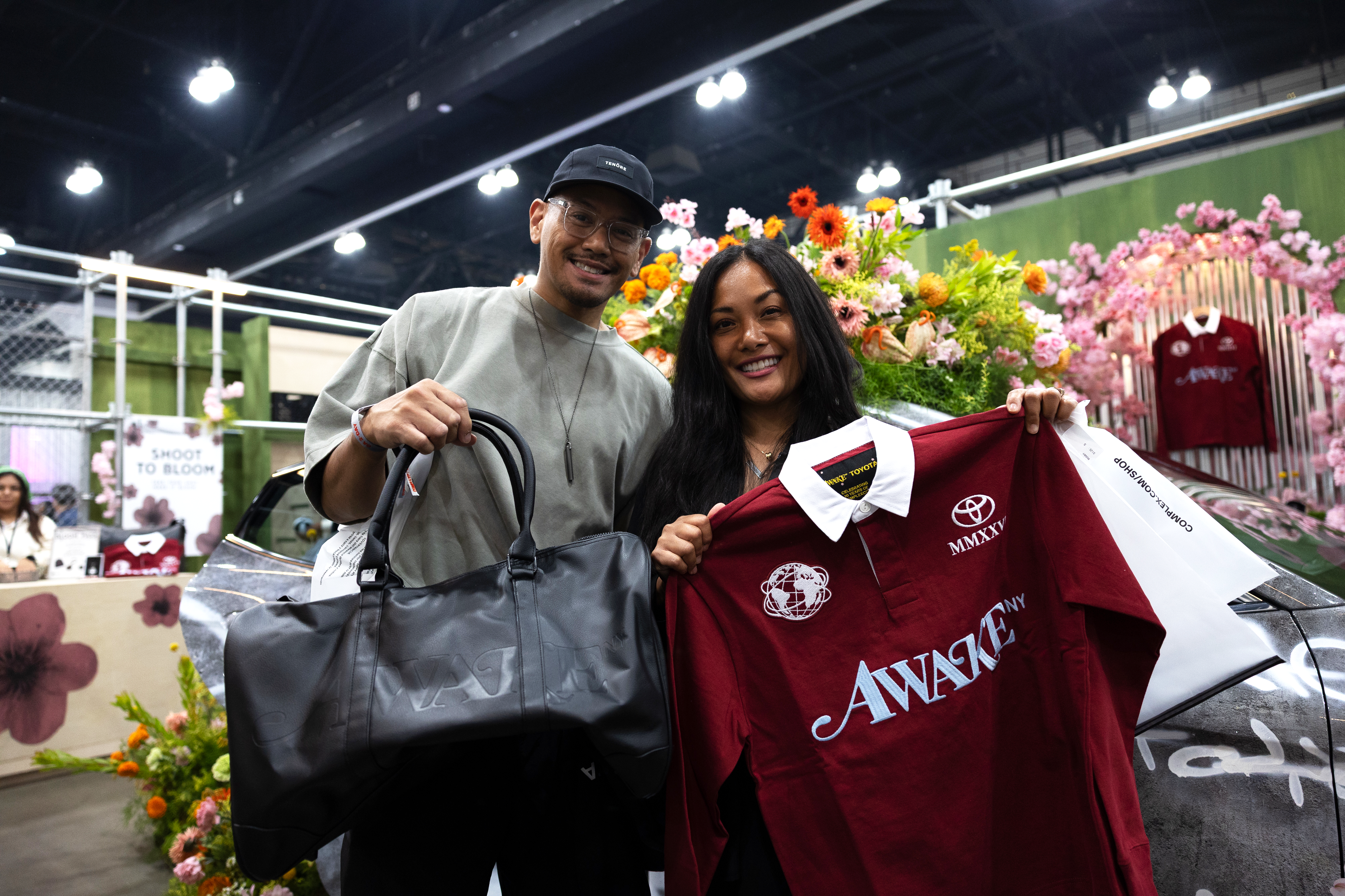 A man and woman smiling at an event, holding a black bag and a maroon "Awake" jersey. The background features colorful flowers.