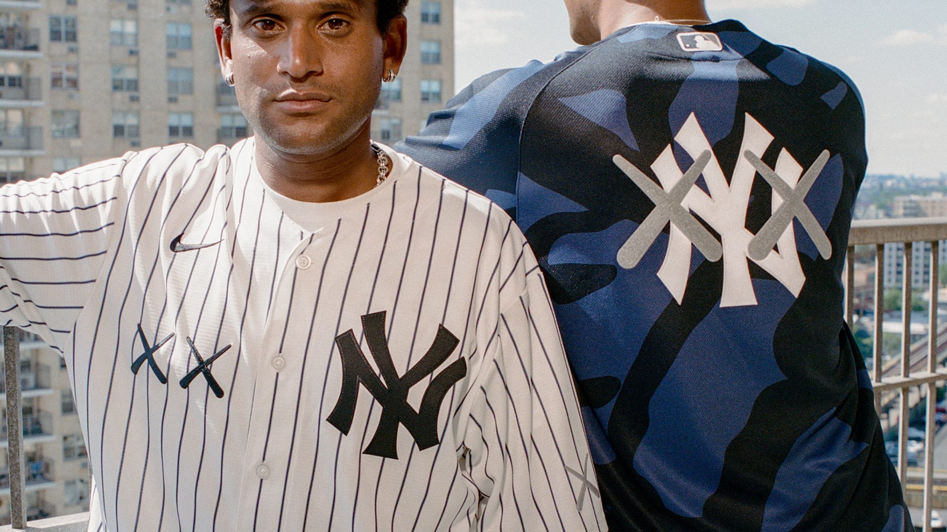 Two people wearing New York Yankees apparel stand on a balcony. One faces forward in a pinstripe jersey, the other faces away in a camo shirt.