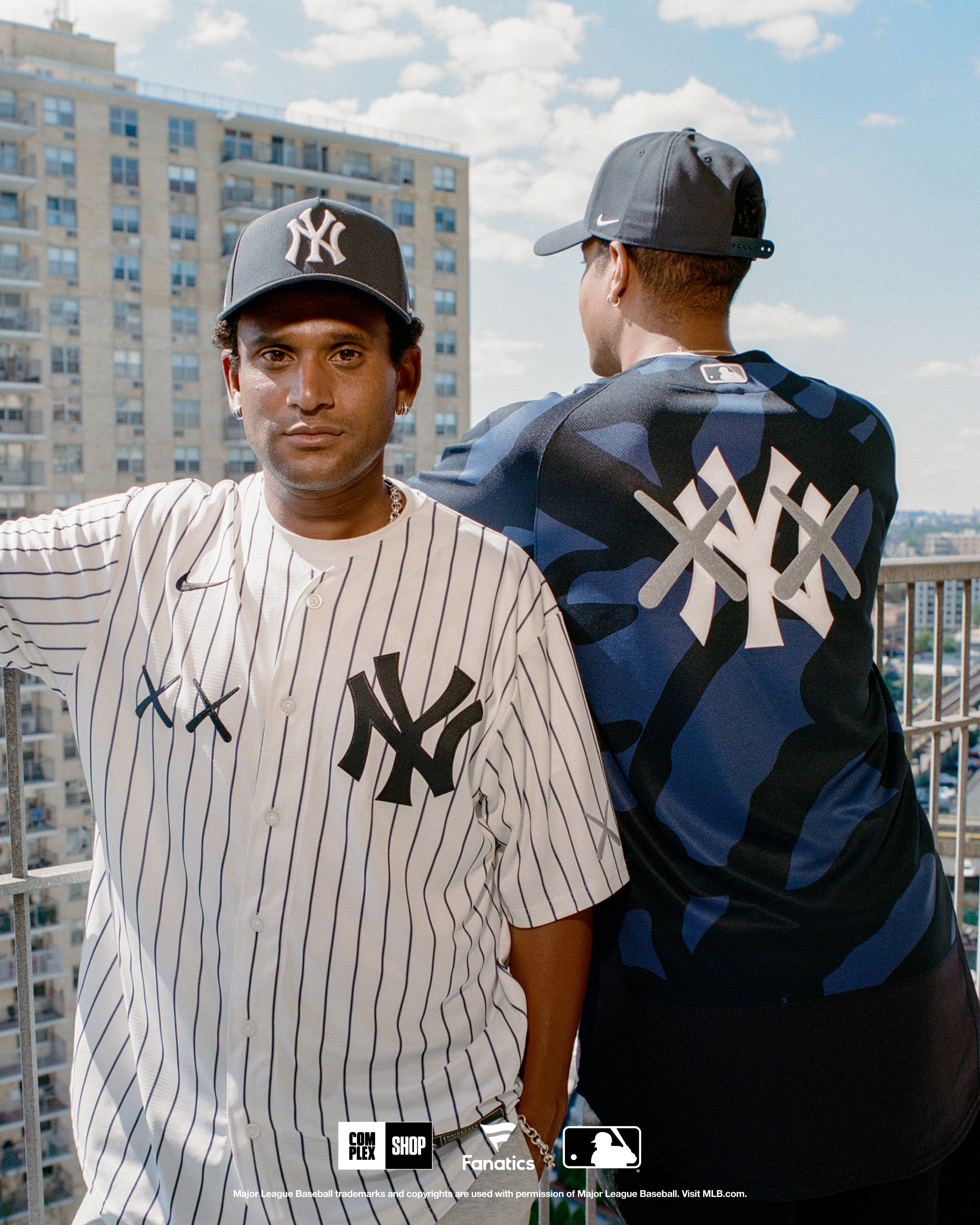 Two people wearing New York Yankees apparel stand on a balcony. One faces forward in a pinstripe jersey, the other faces away in a camo shirt.
