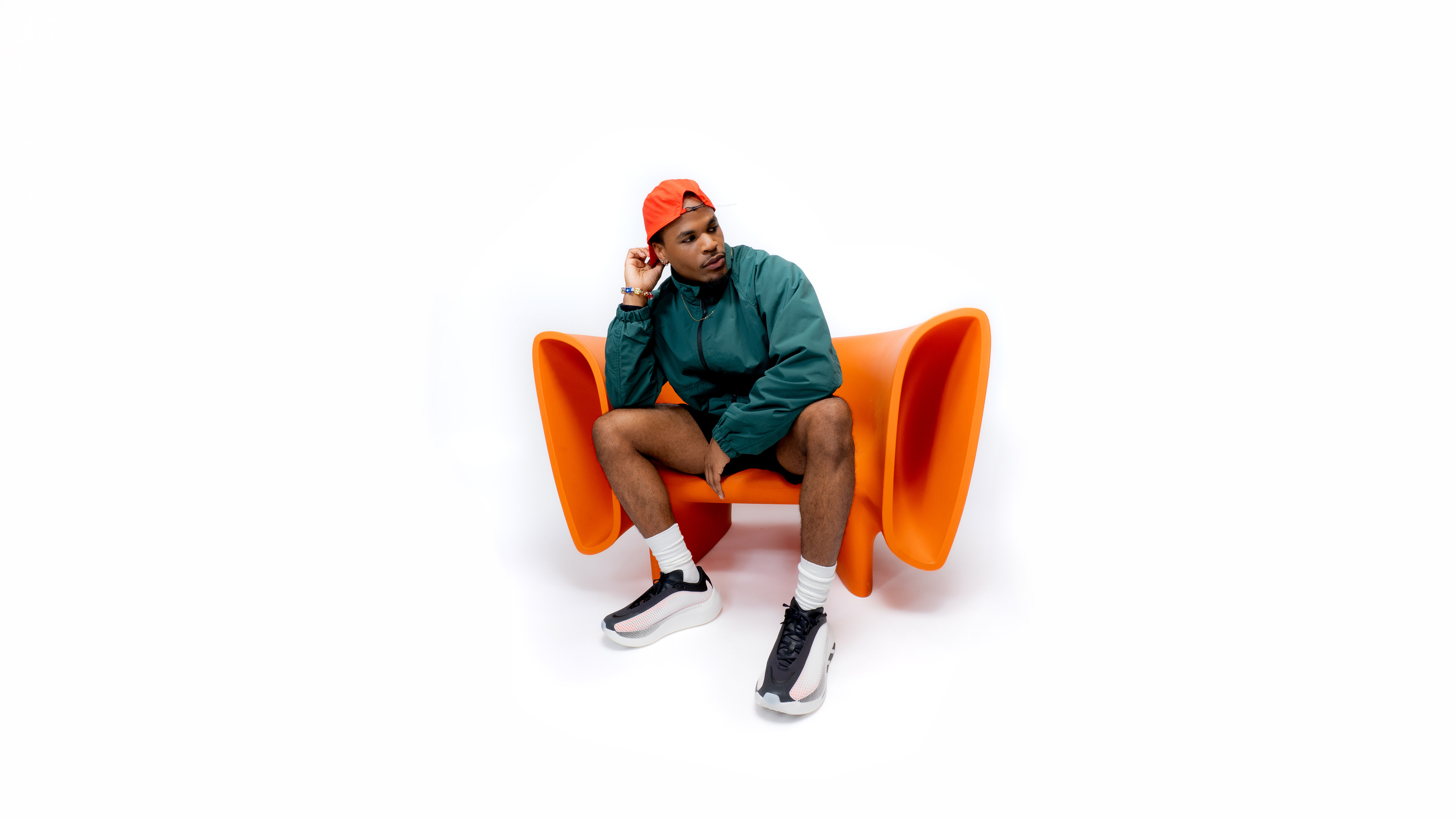 A person in a green jacket and red cap sits on an orange chair, holding a typewriter. They wear black shorts and sneakers.