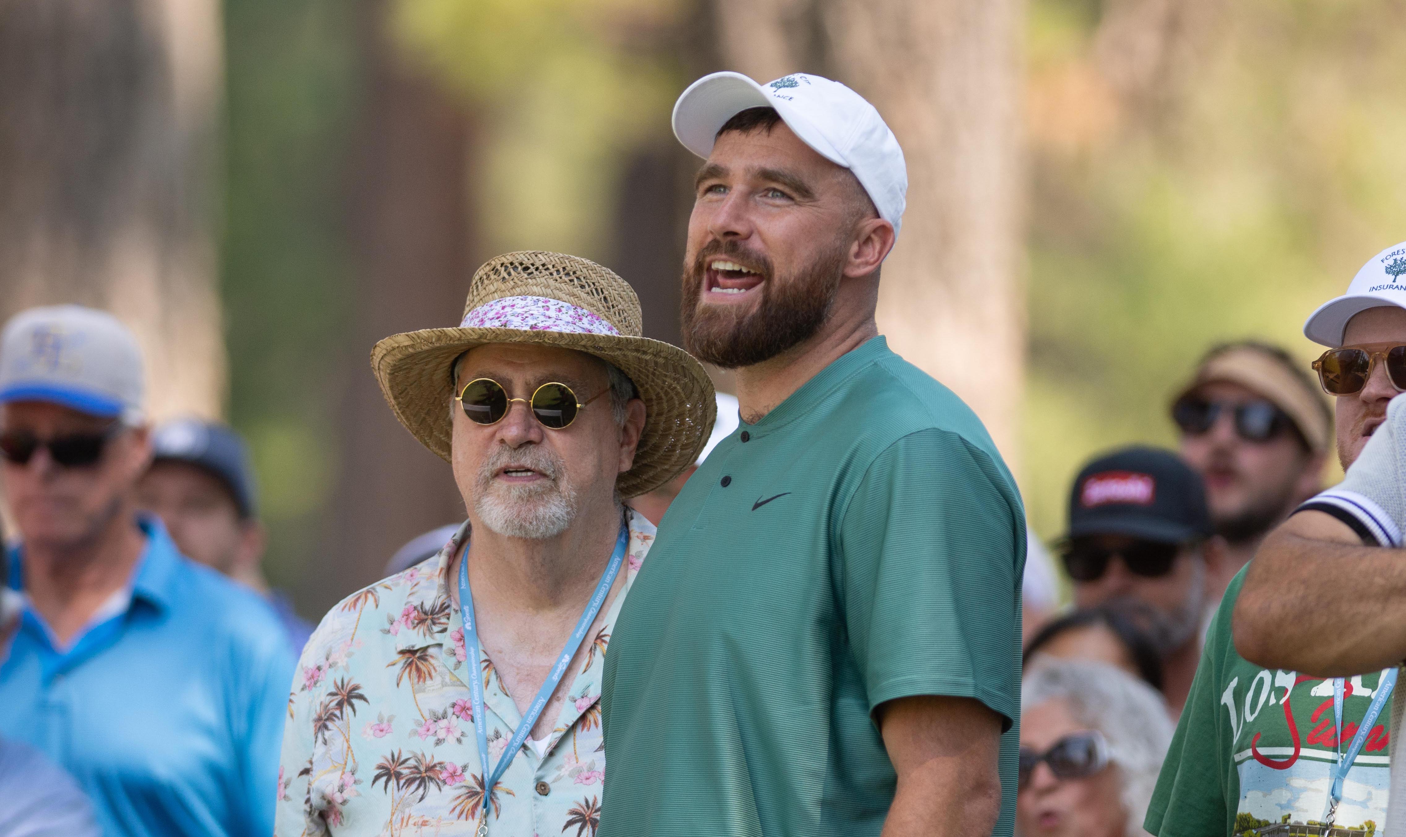 Ed Kelce wears a straw hat and sunglasses stands next to a man in a white cap and green shirt, both looking upwards, surrounded by a crowd.