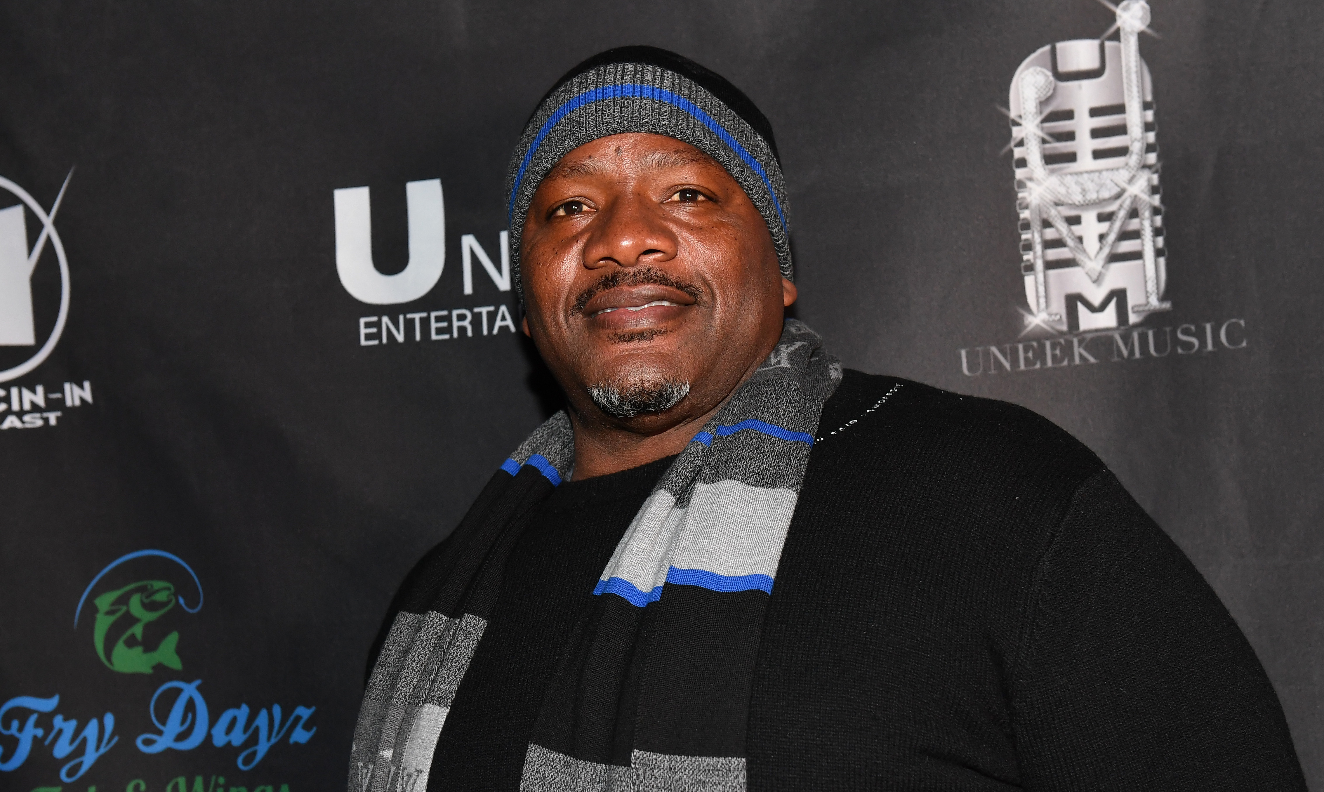 Eugene "Big U" Henley wearing a black sweater, gray and blue scarf, and a beanie stands in front of a backdrop with logos.