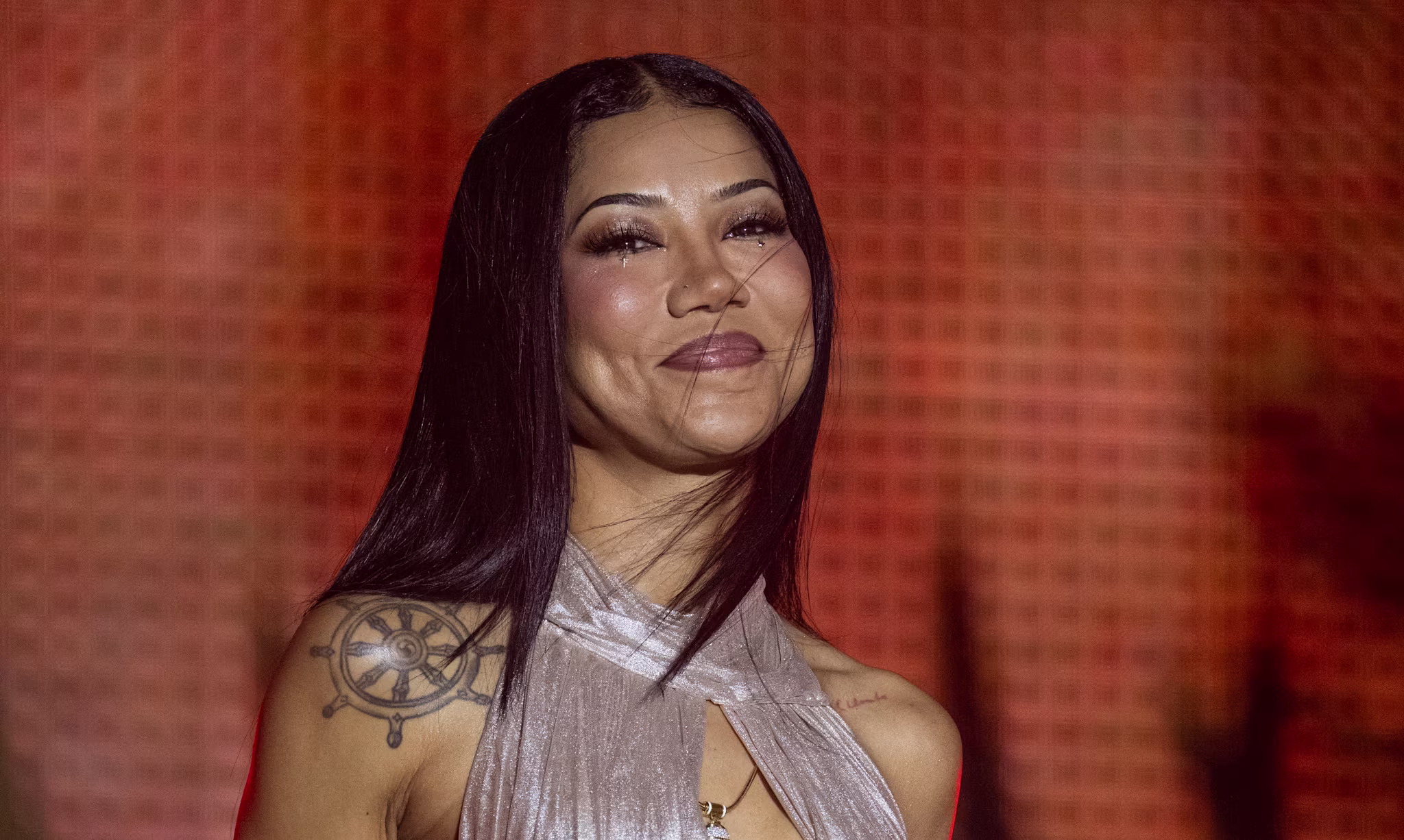 Jhene Aiko in a shimmering silver dress with tattoos stands against a warm, textured background, surrounded by palm leaves.