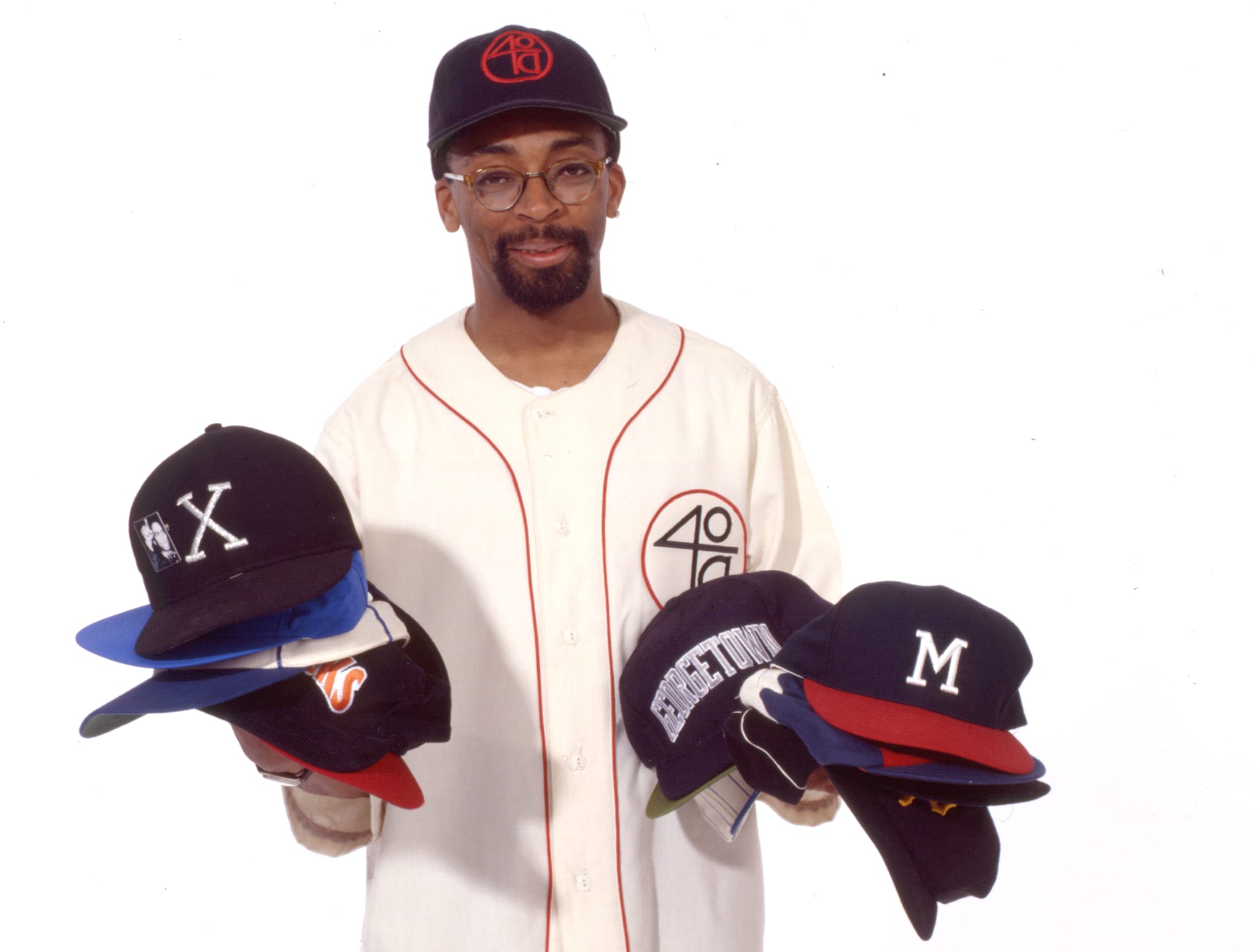 A man wearing a baseball jersey and cap, holding multiple caps in each hand.