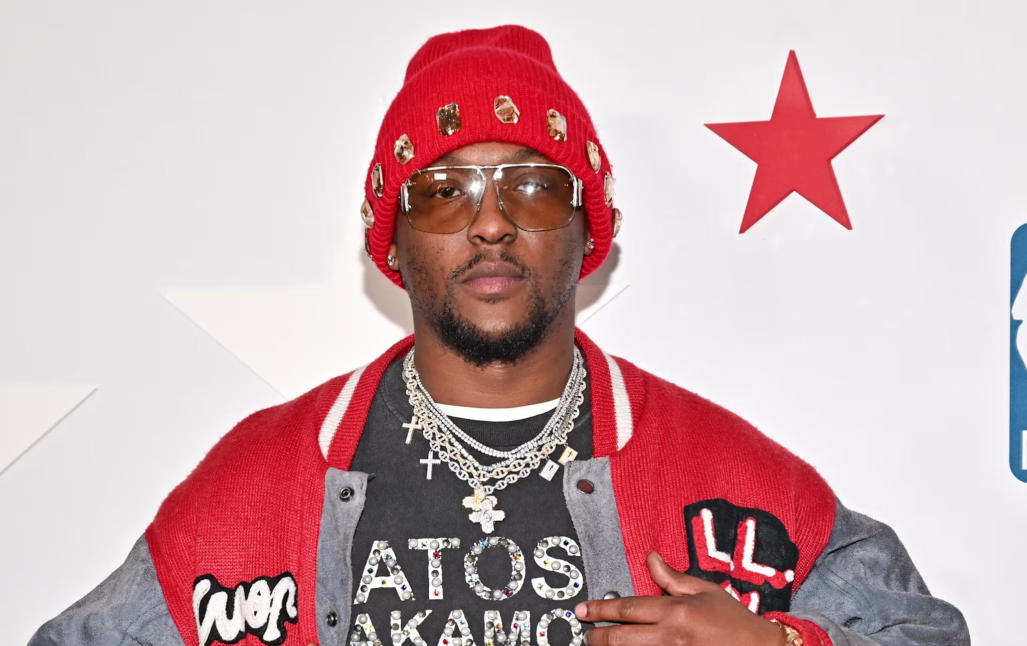 Hit-Boy wearing a red beanie, sunglasses, a red and gray jacket, and a black shirt with chains, poses at an event.