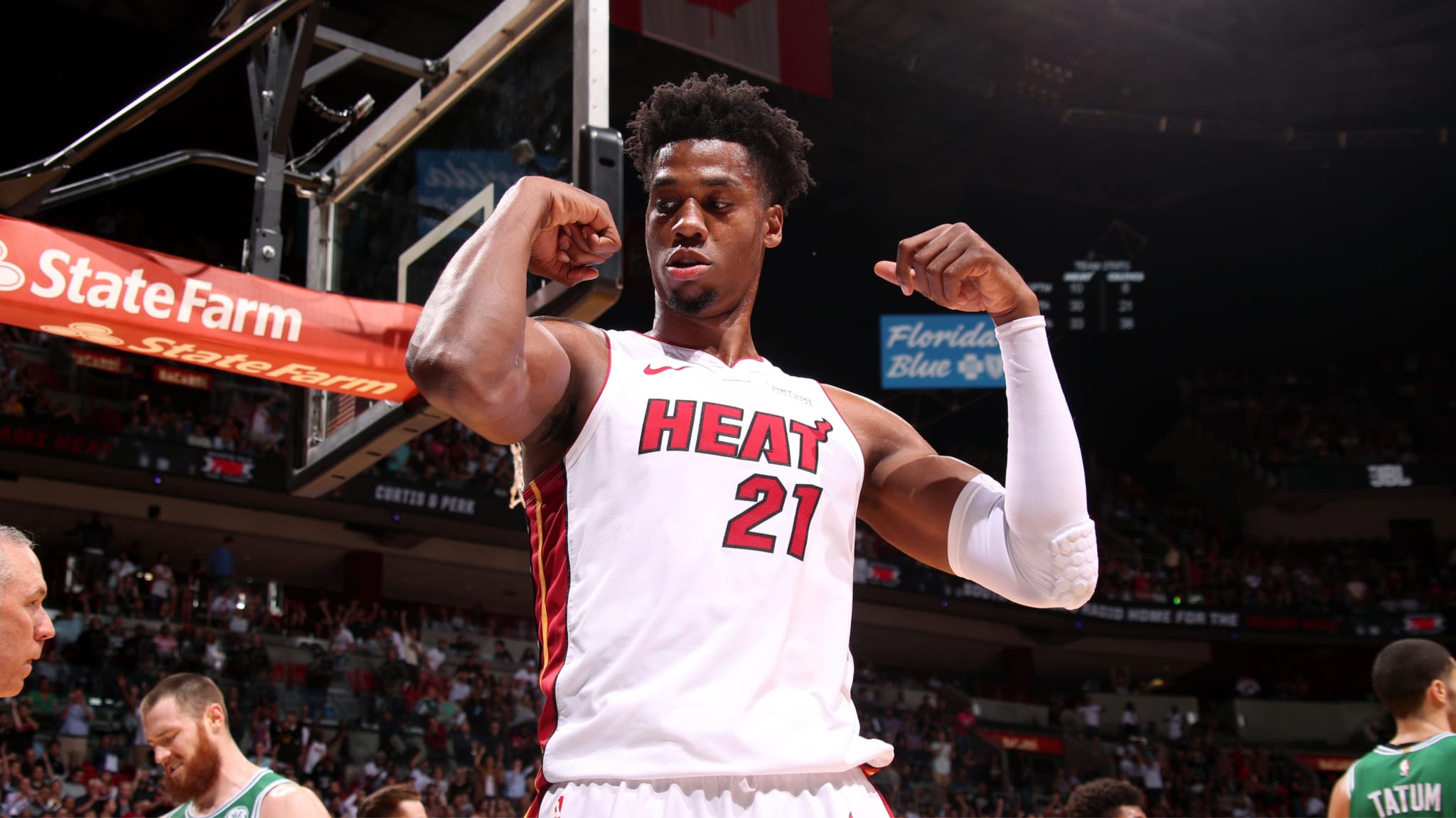 Hassan Whiteside in a Miami Heat jersey flexes his muscles on the court, with spectators and players in the background.