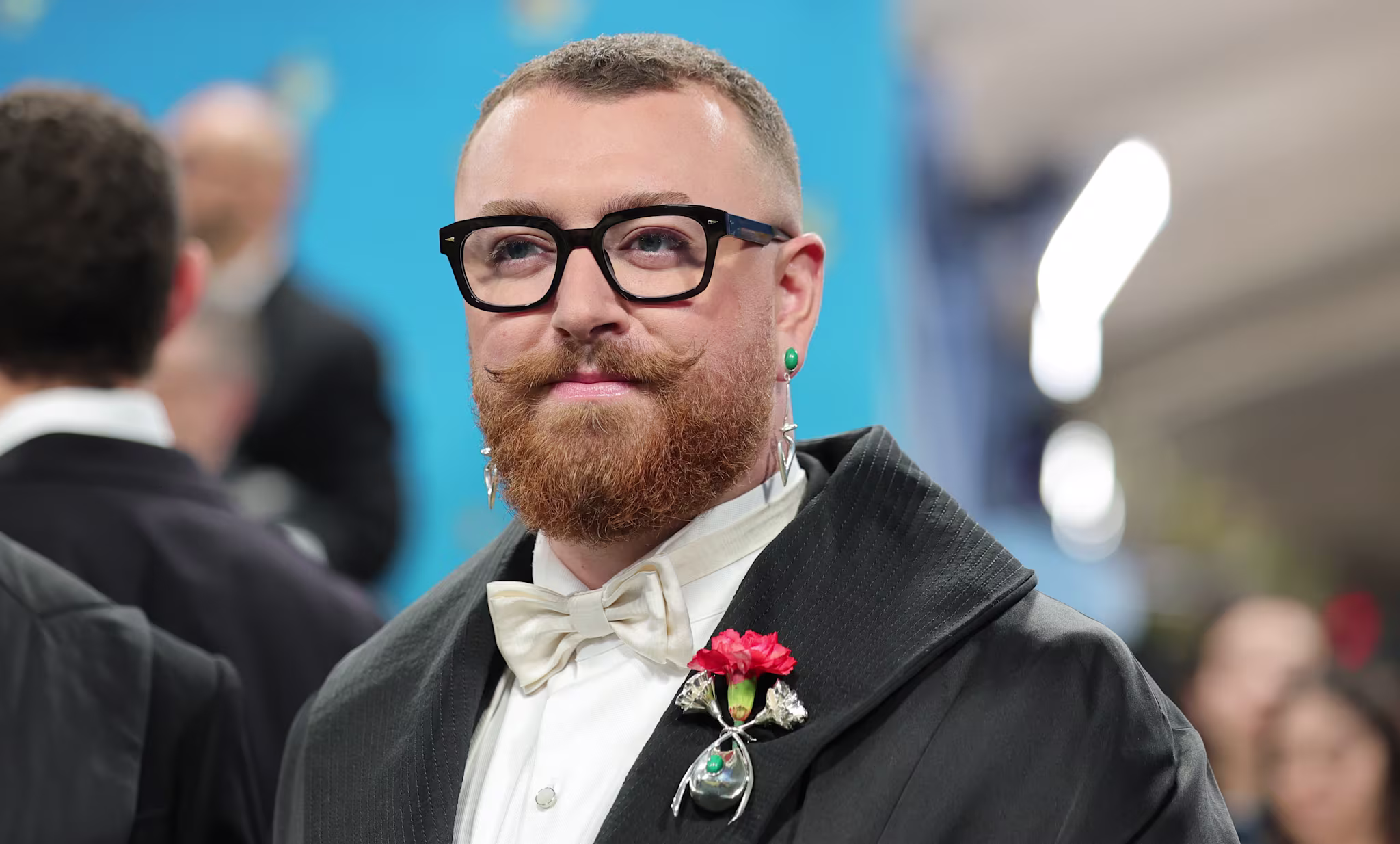 Sam Smith with glasses, a beard, and a formal outfit with a red flower brooch at a formal event.