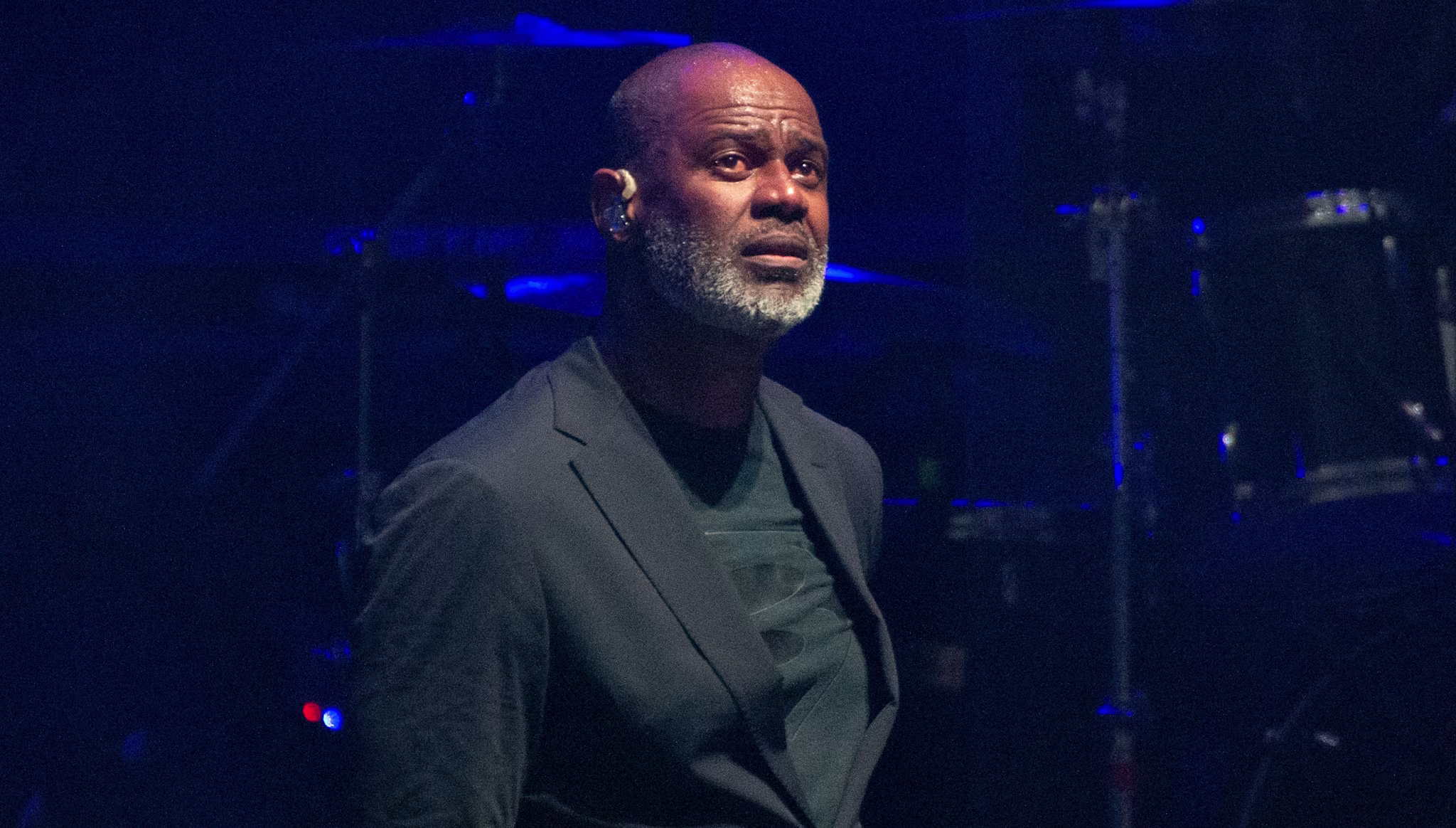 Brian McKnight with a beard in a dark suit stands on stage, with drum equipment in the background.